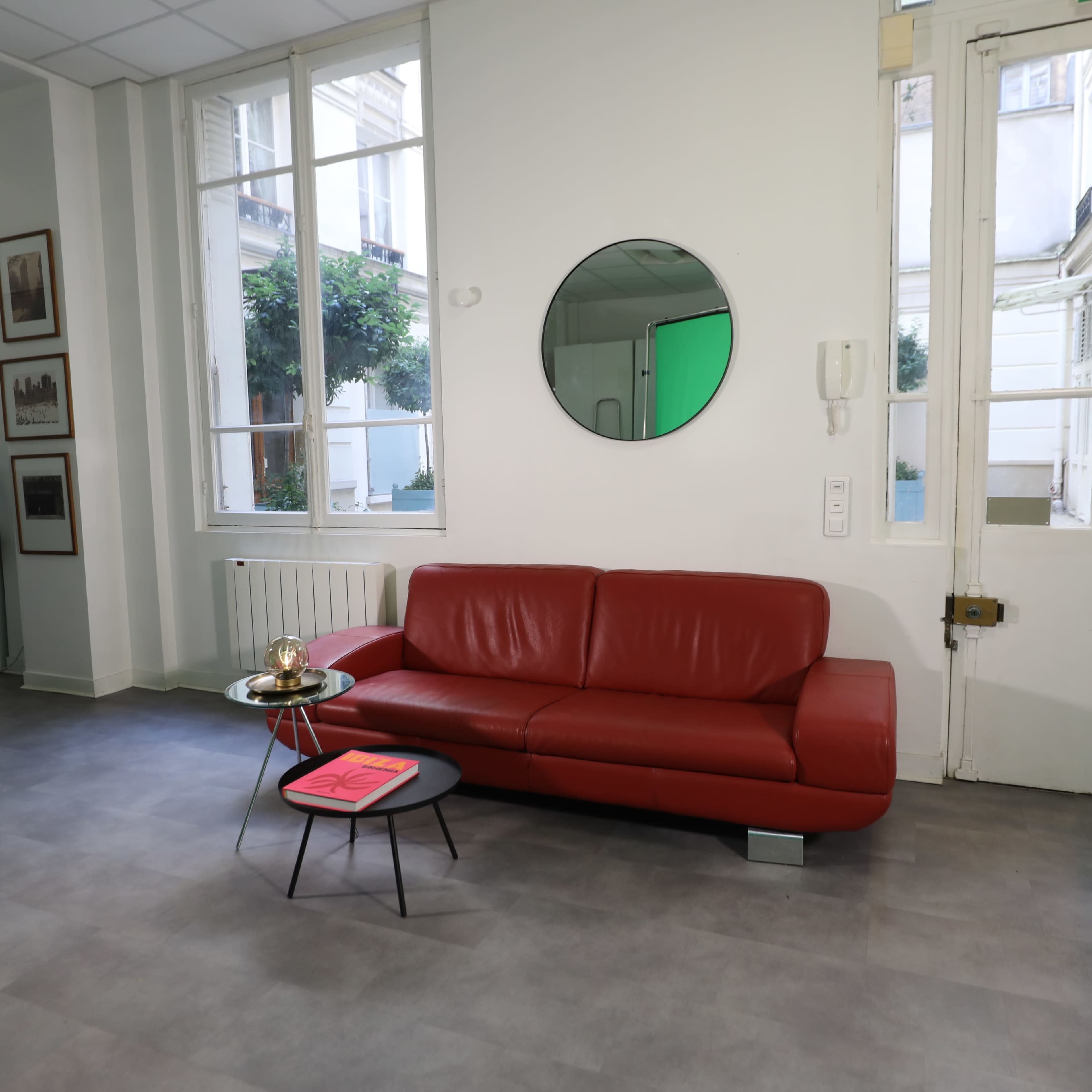 A modern interior with a red couch, a small black table, and a mirror on the wall, illuminated by natural light from large windows.