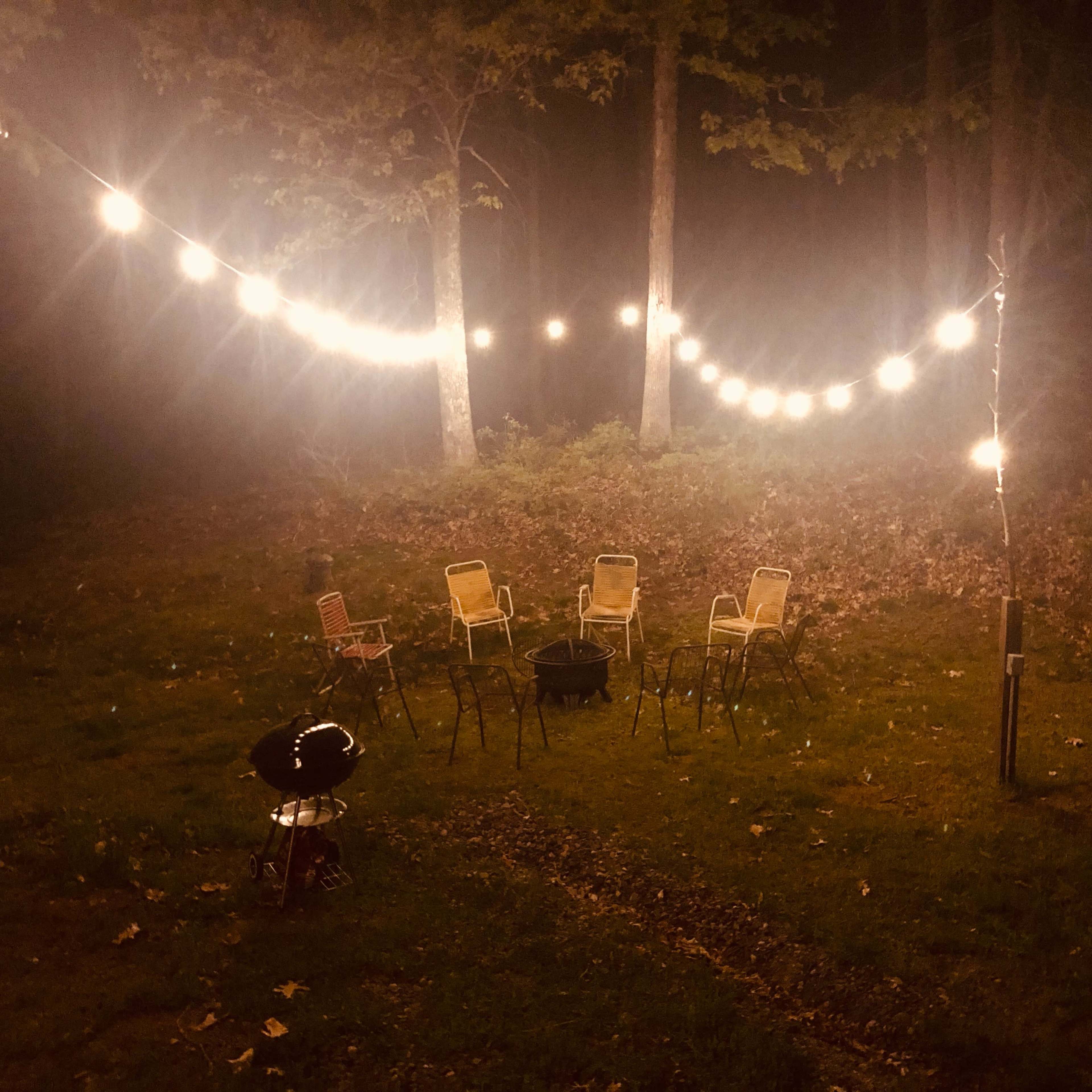 A circle of empty chairs is arranged around a fire pit, illuminated by string lights in a forest setting at night.