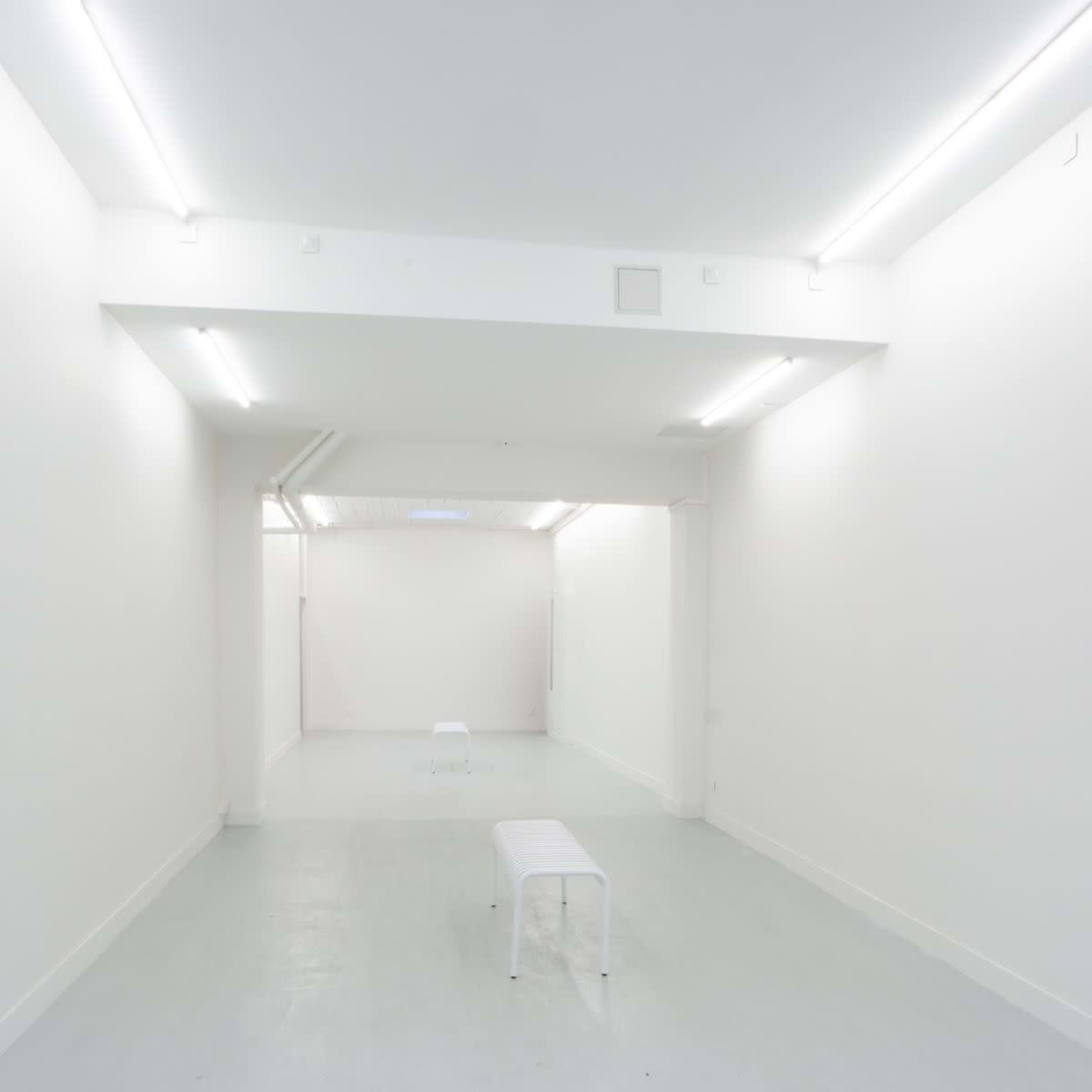 A minimalistic, brightly lit white room featuring two white benches positioned on a light-colored floor.