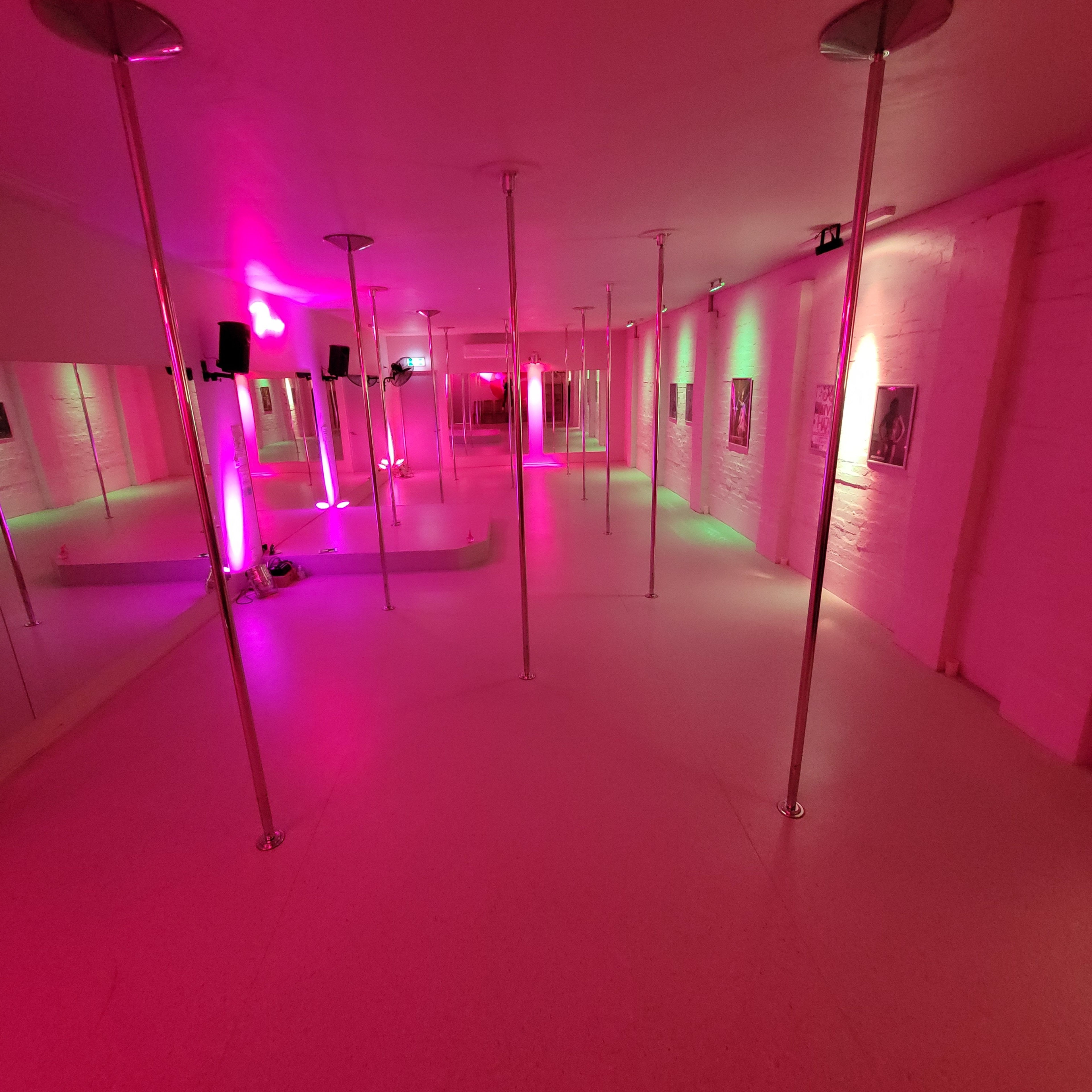 A spacious dance studio featuring multiple mirrored walls and several polished dance poles illuminated by pink and green lights.