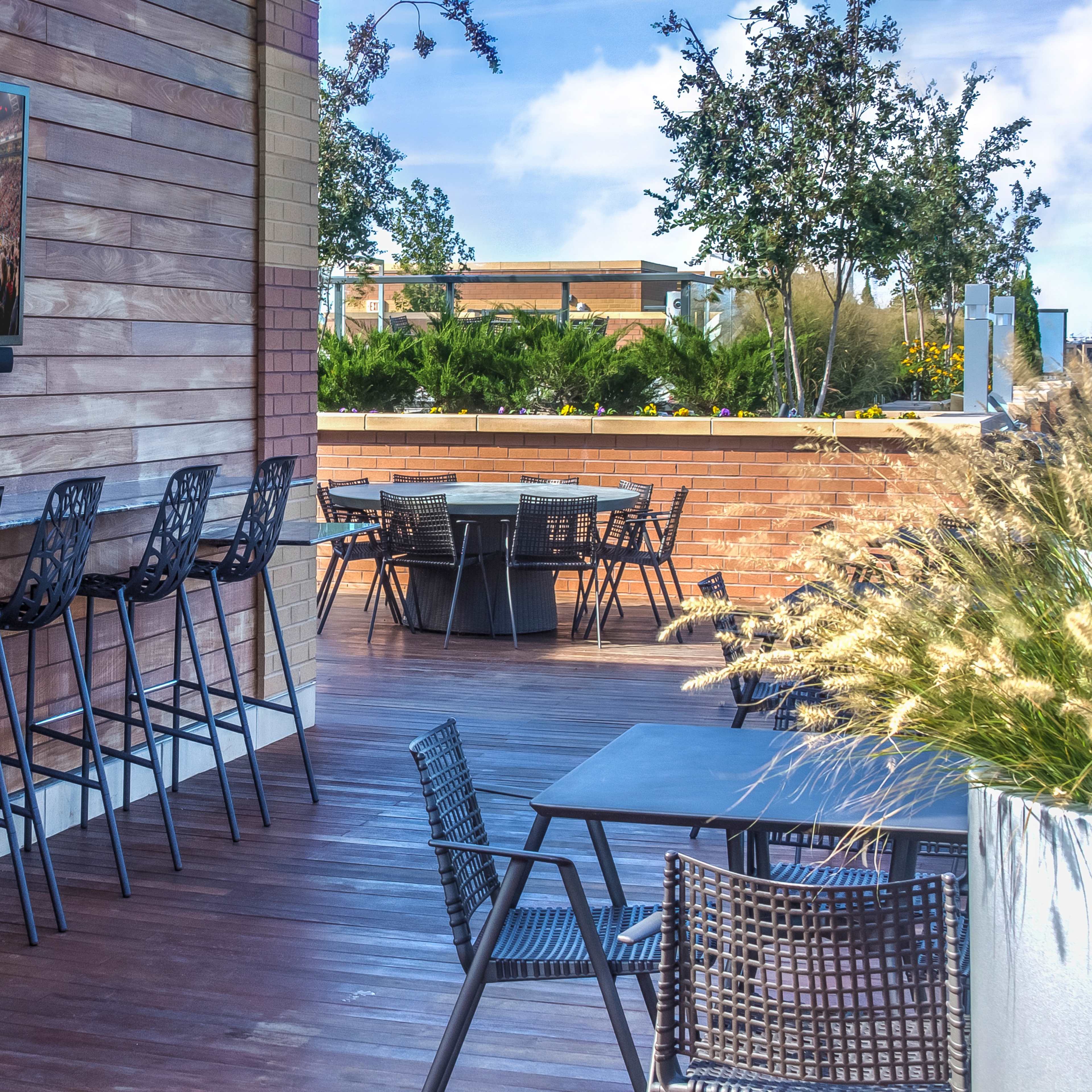 A modern outdoor seating area with wooden walls, high bar stools, a flat-screen television mounted on the wall, and tables arranged on a wooden deck surrounded by greenery.