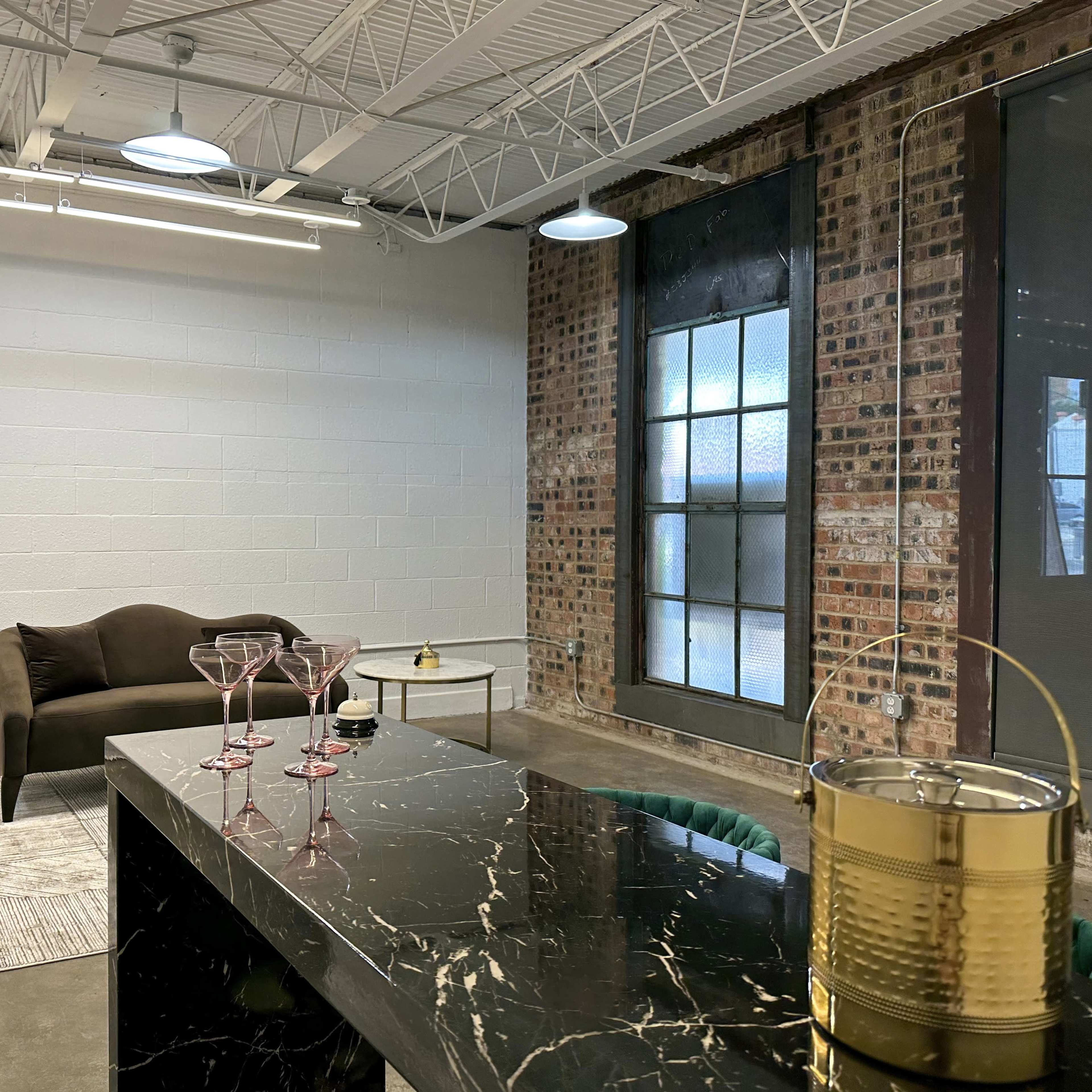 A modern office or lounge space with a dark sofa, a glass coffee table, a patterned rug, and a bar cart with glasses and a decorative item.