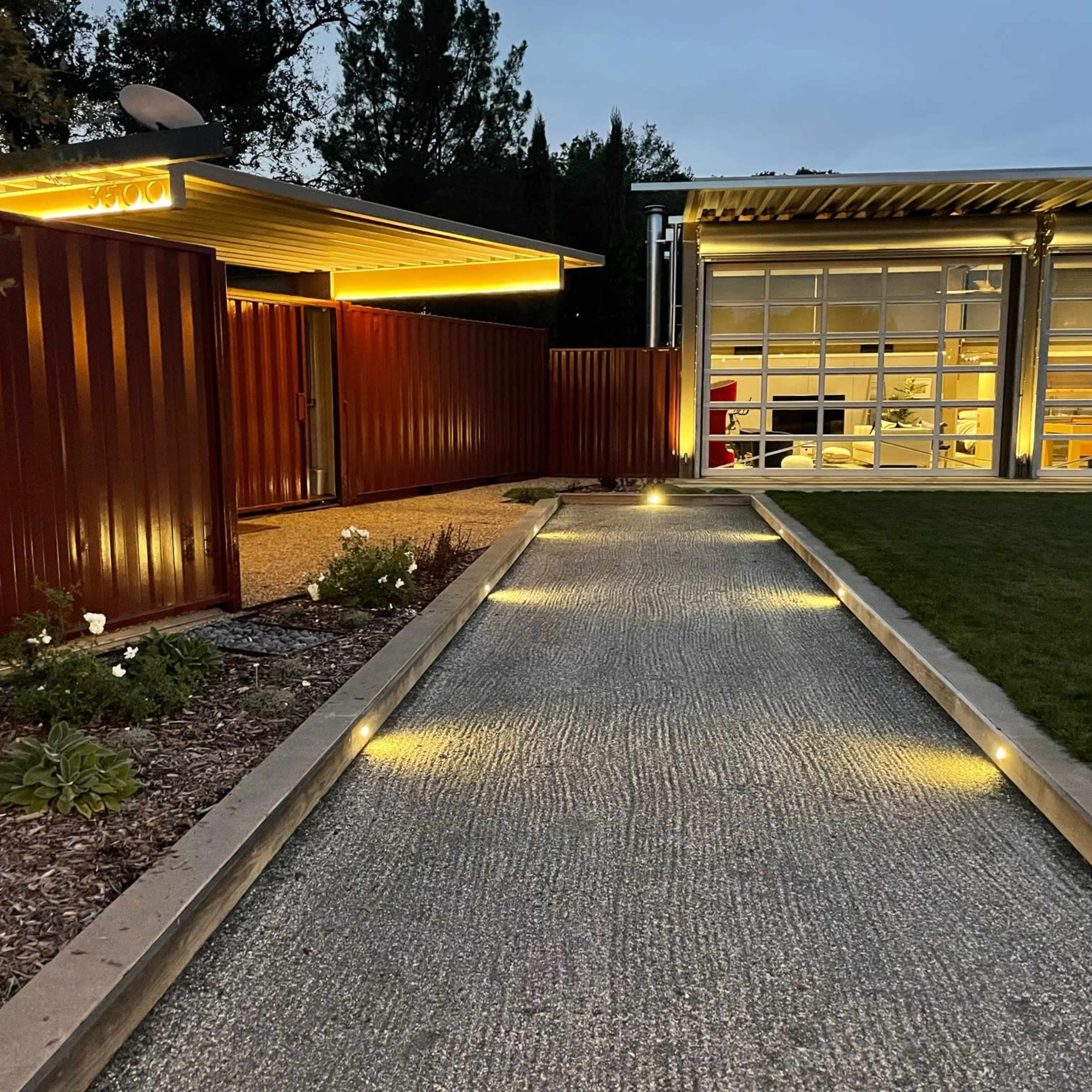 A gravel pathway lined with lights leads to a modern building with a glass garage door and a fenced entryway.