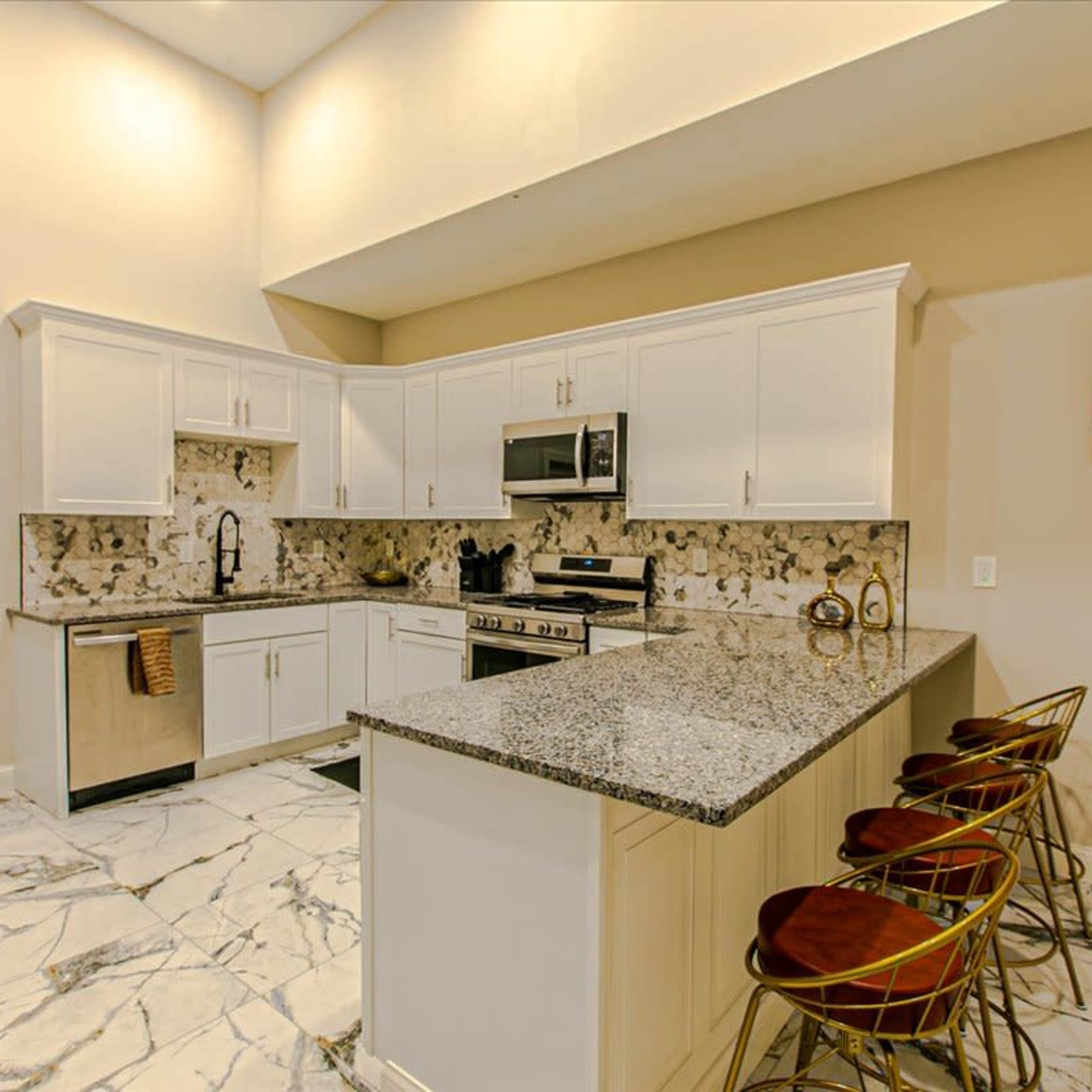 A modern kitchen featuring white cabinetry, stainless steel appliances, and a granite island with three gold bar stools.
