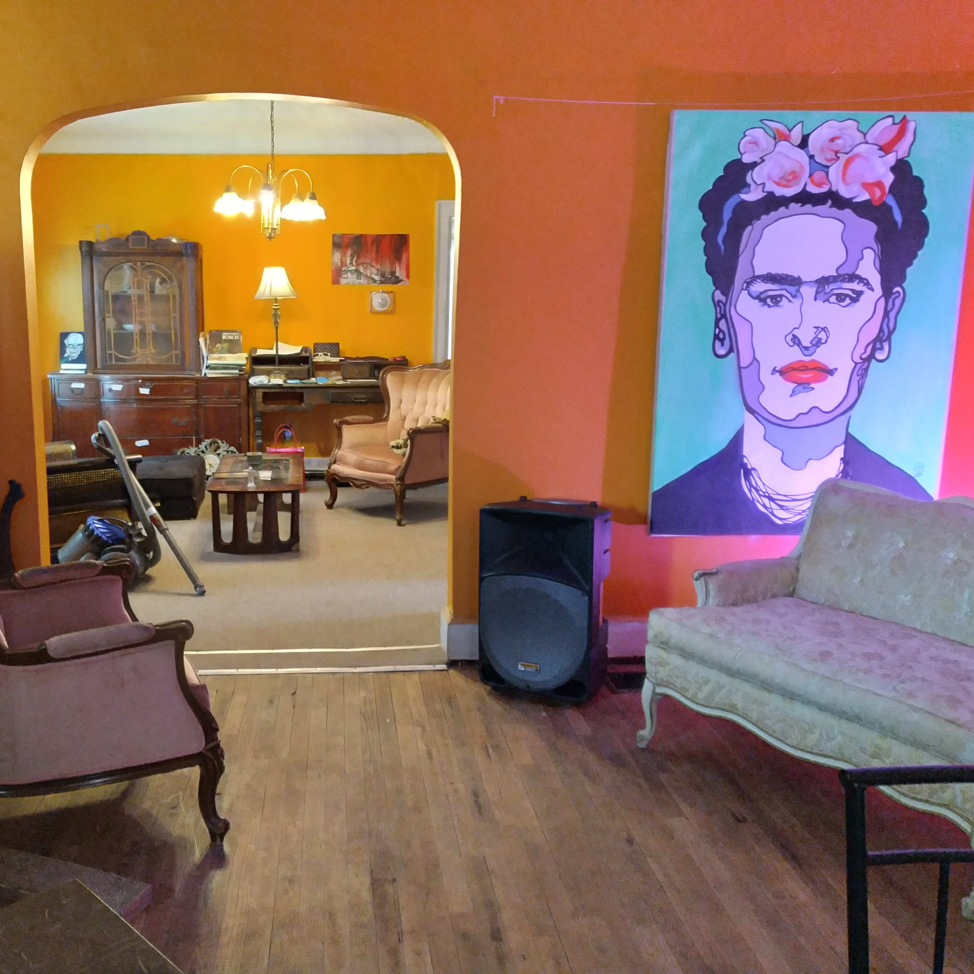 A brightly painted orange room with a vintage decor featuring two armchairs, a small coffee table, a refrigerator covered in stickers, and a colorful portrait of Frida Kahlo on the wall.