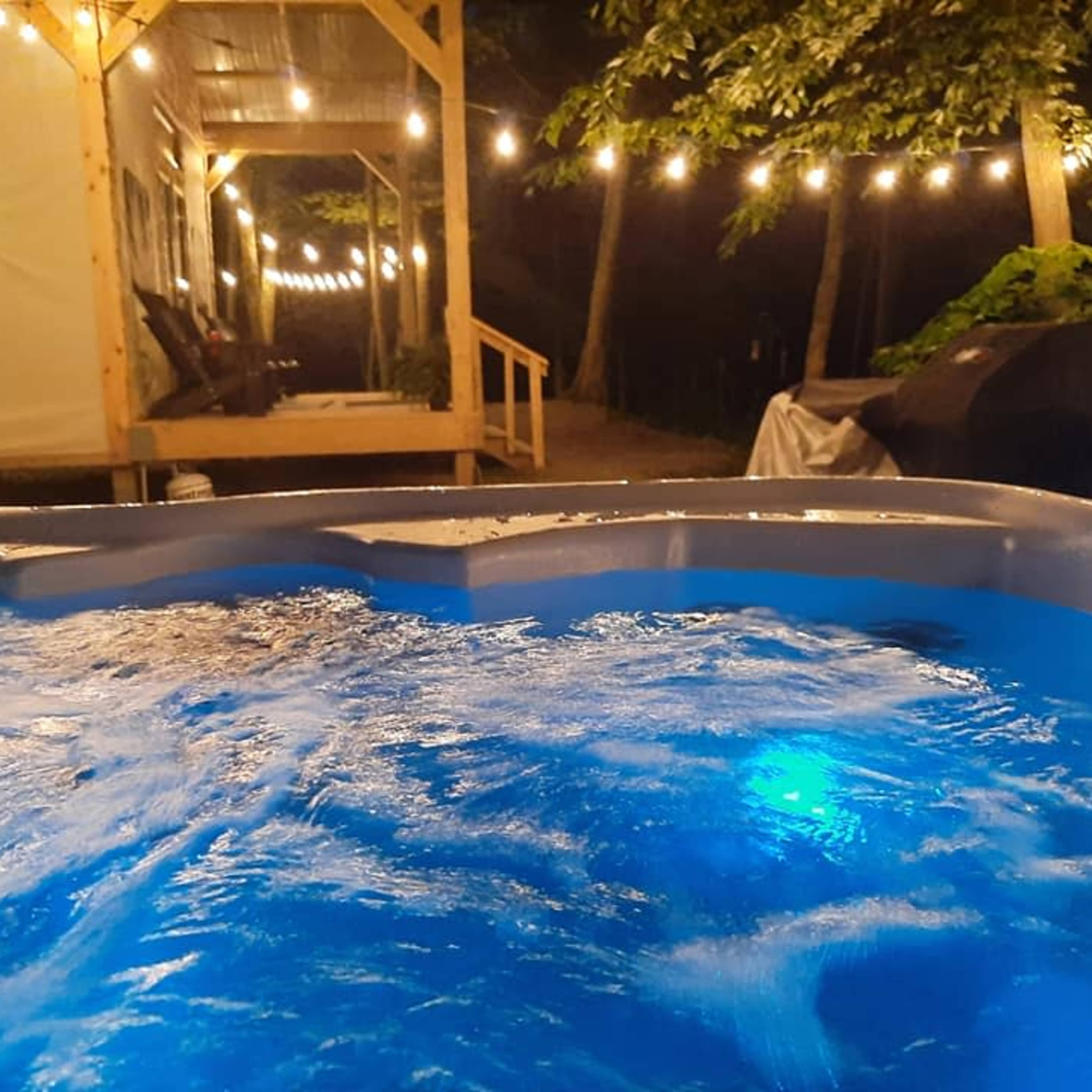 A hot tub illuminated with blue lights, situated near a wooden deck adorned with string lights in a wooded outdoor area at night.