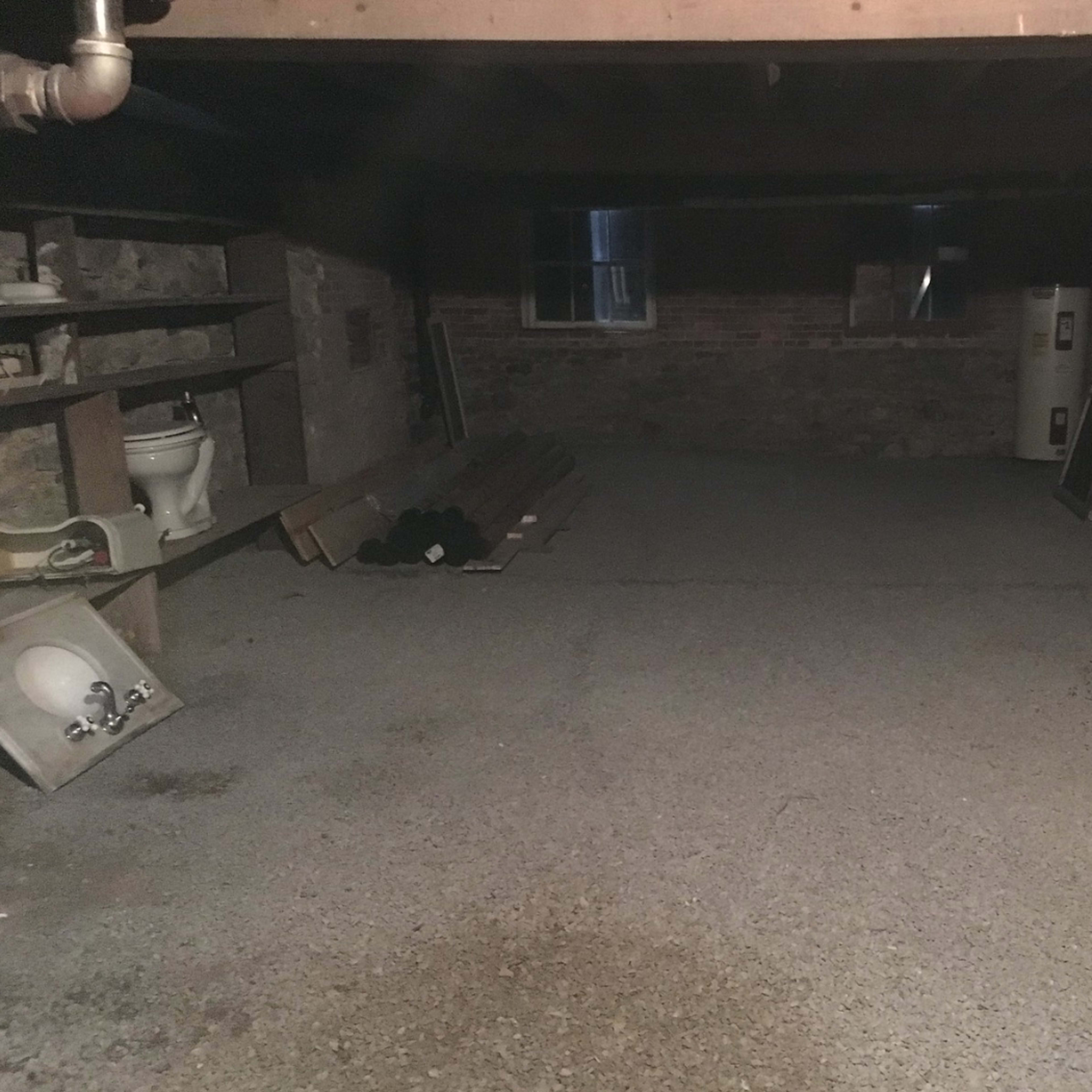 A dimly lit basement with a concrete floor, shelves on one side, and various construction materials stacked against the walls.