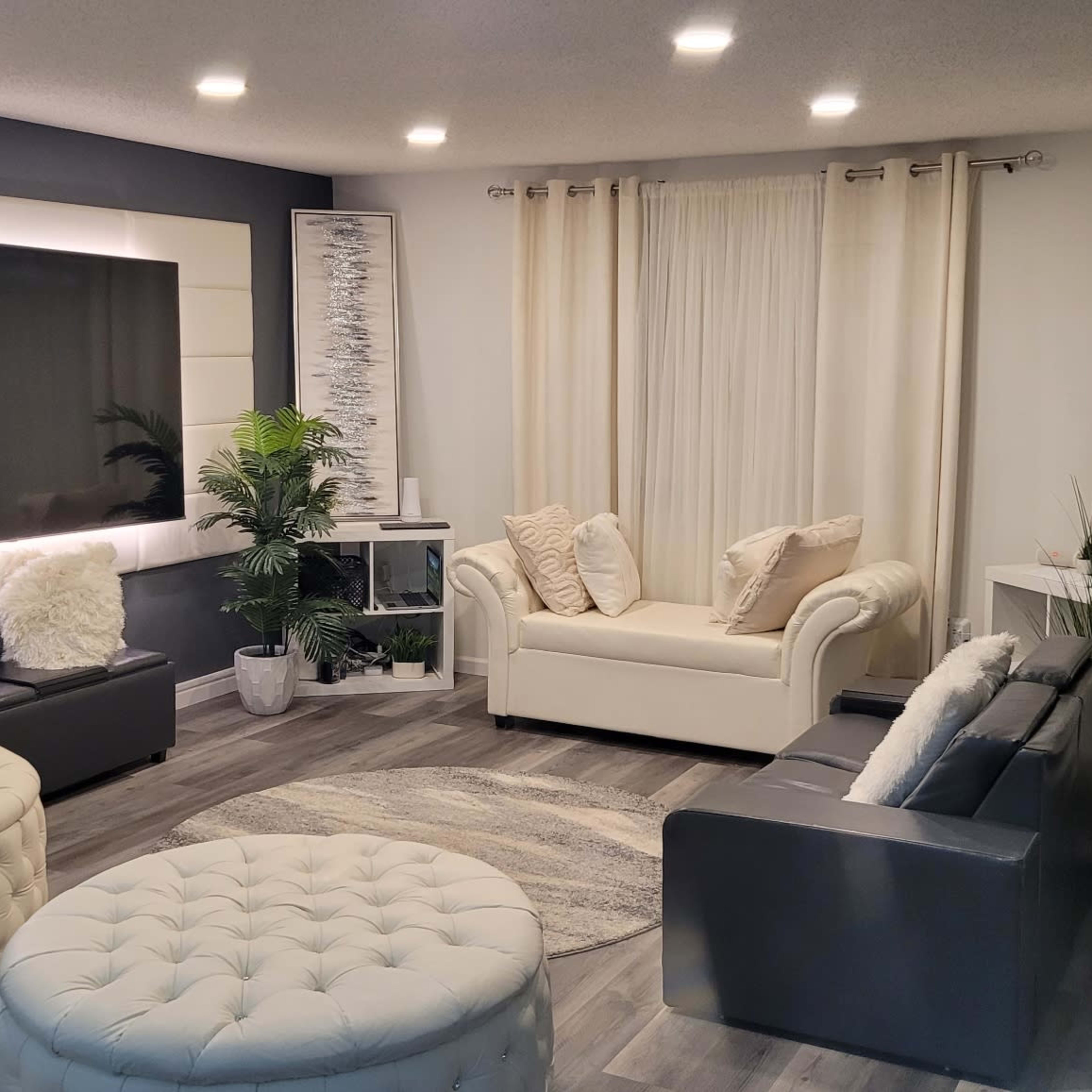 A modern living room with white sofas, round ottomans, and a large television against a feature wall, complemented by plants and curtains.