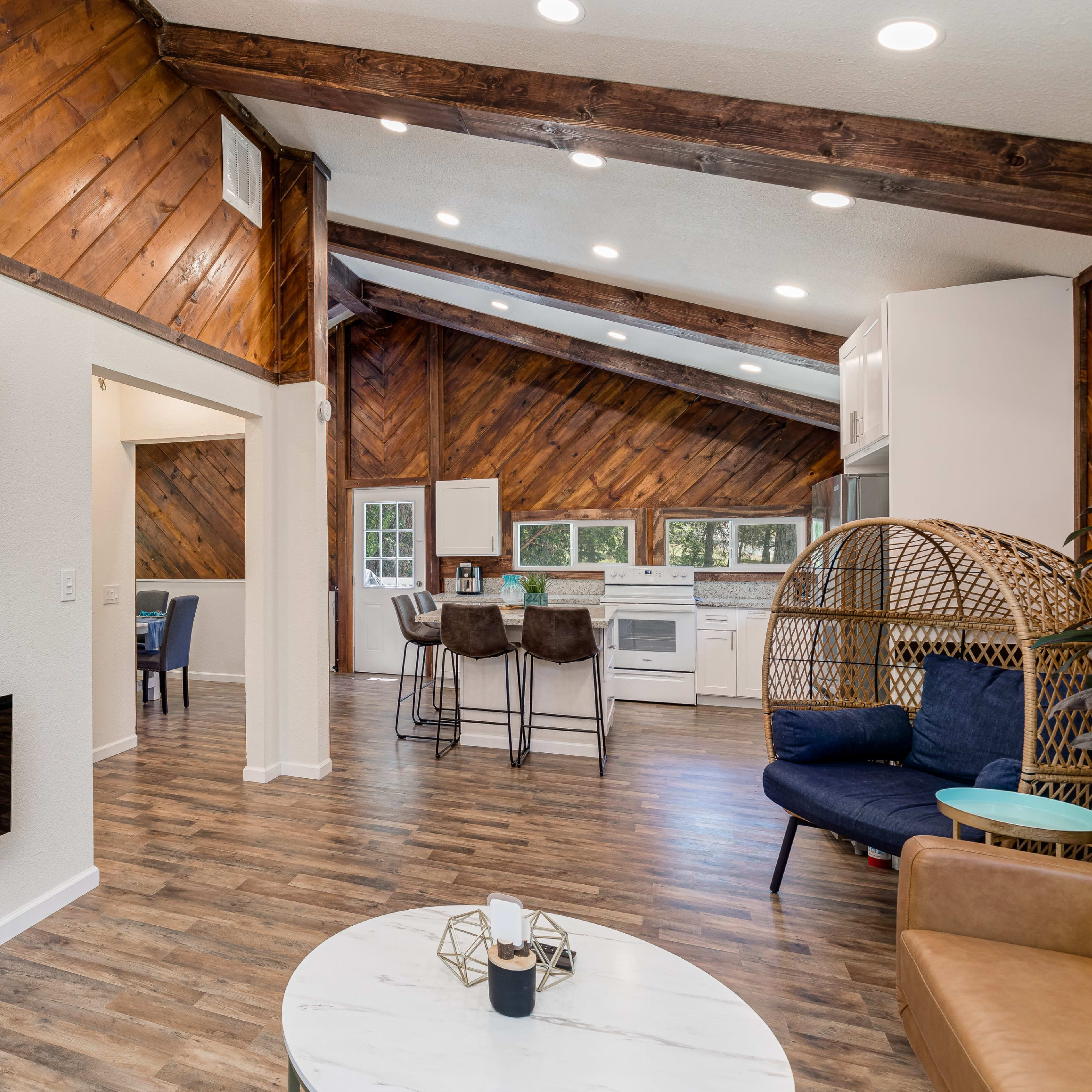 A modern living room with a flat-screen TV mounted on the wall, a contemporary electric fireplace, and an open kitchen area featuring white cabinetry and a wooden ceiling.