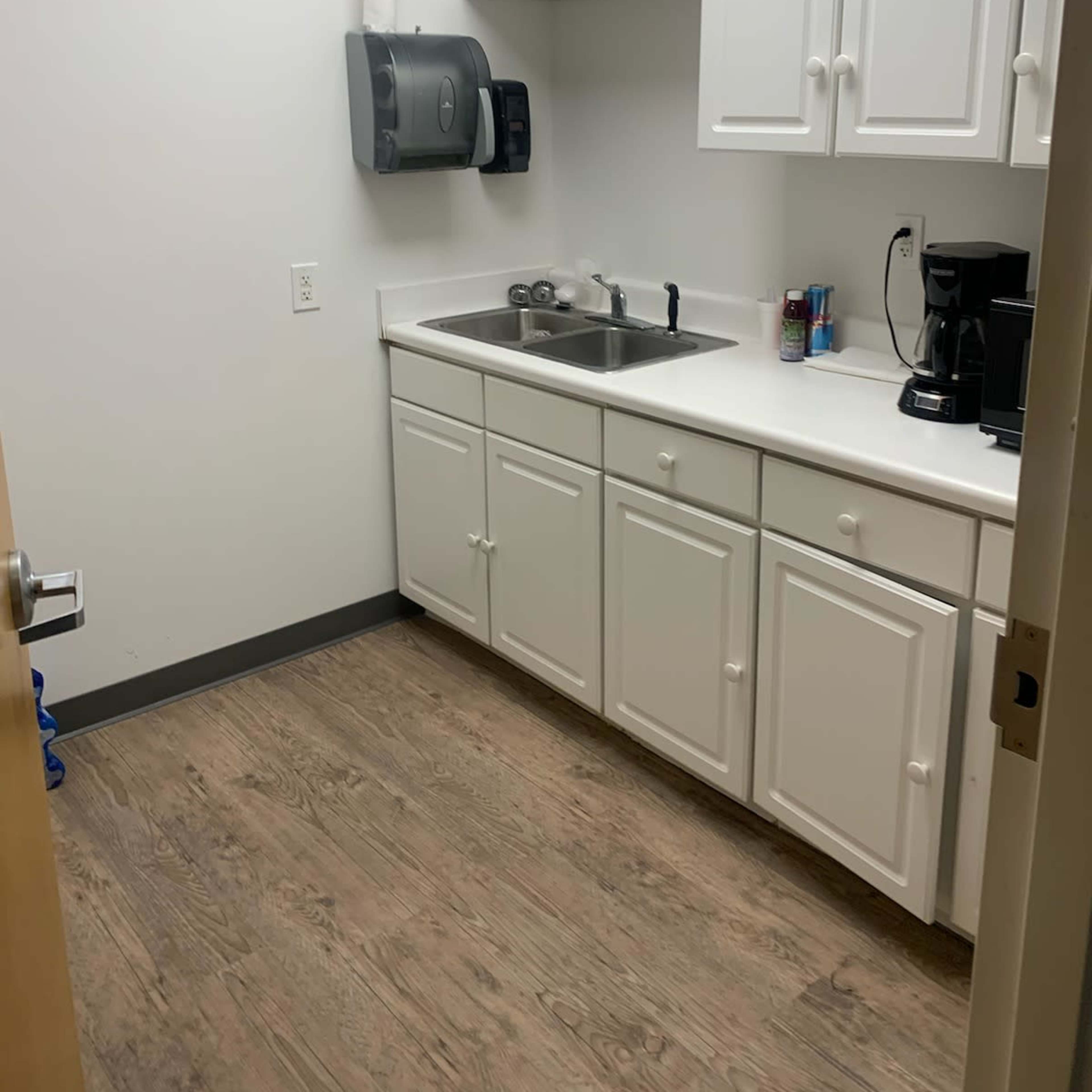 A small kitchenette with white cabinets, a sink, and a coffee maker against a plain wall.