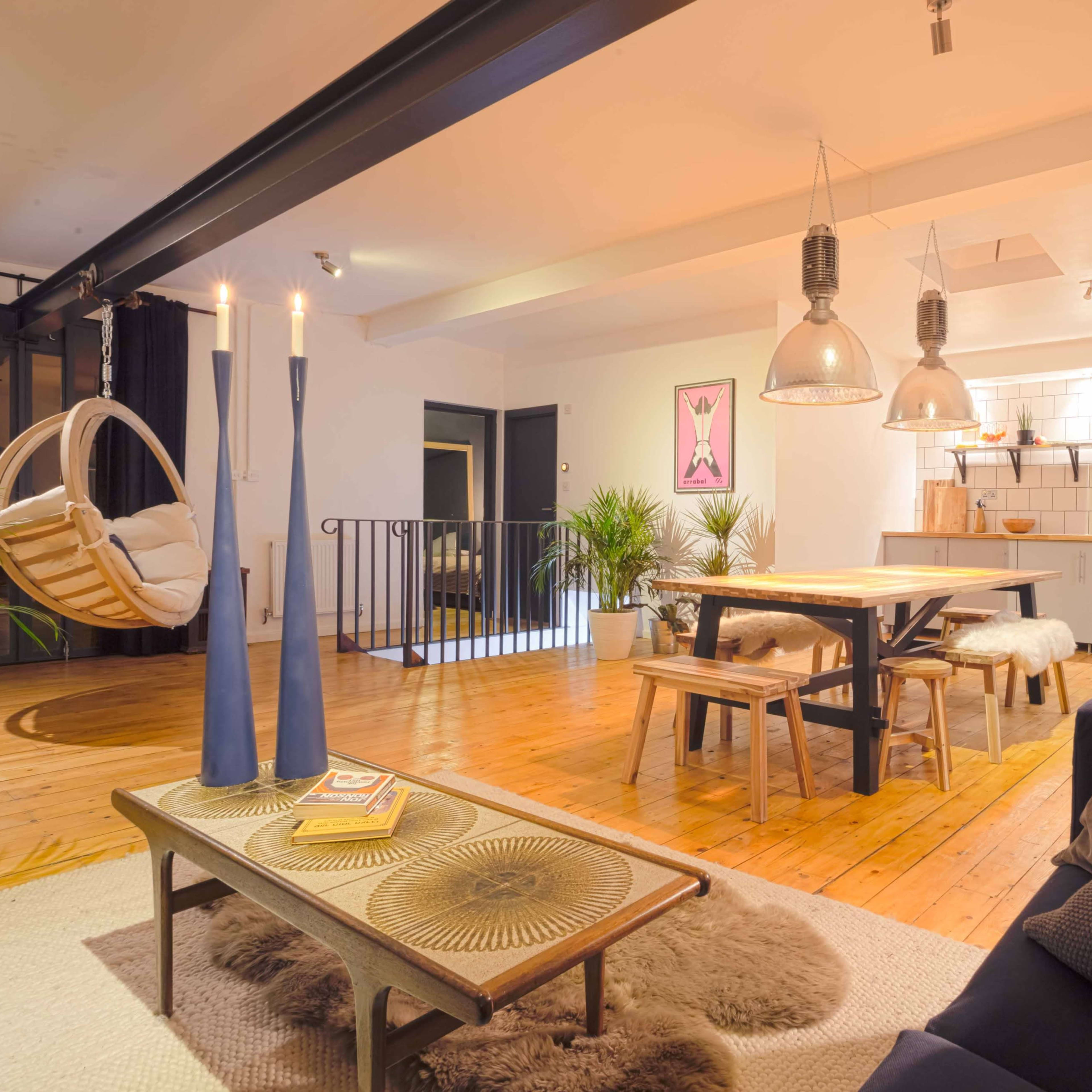 A modern living room featuring a hanging chair, a dining table with benches, and a well-lit kitchen area in the background.