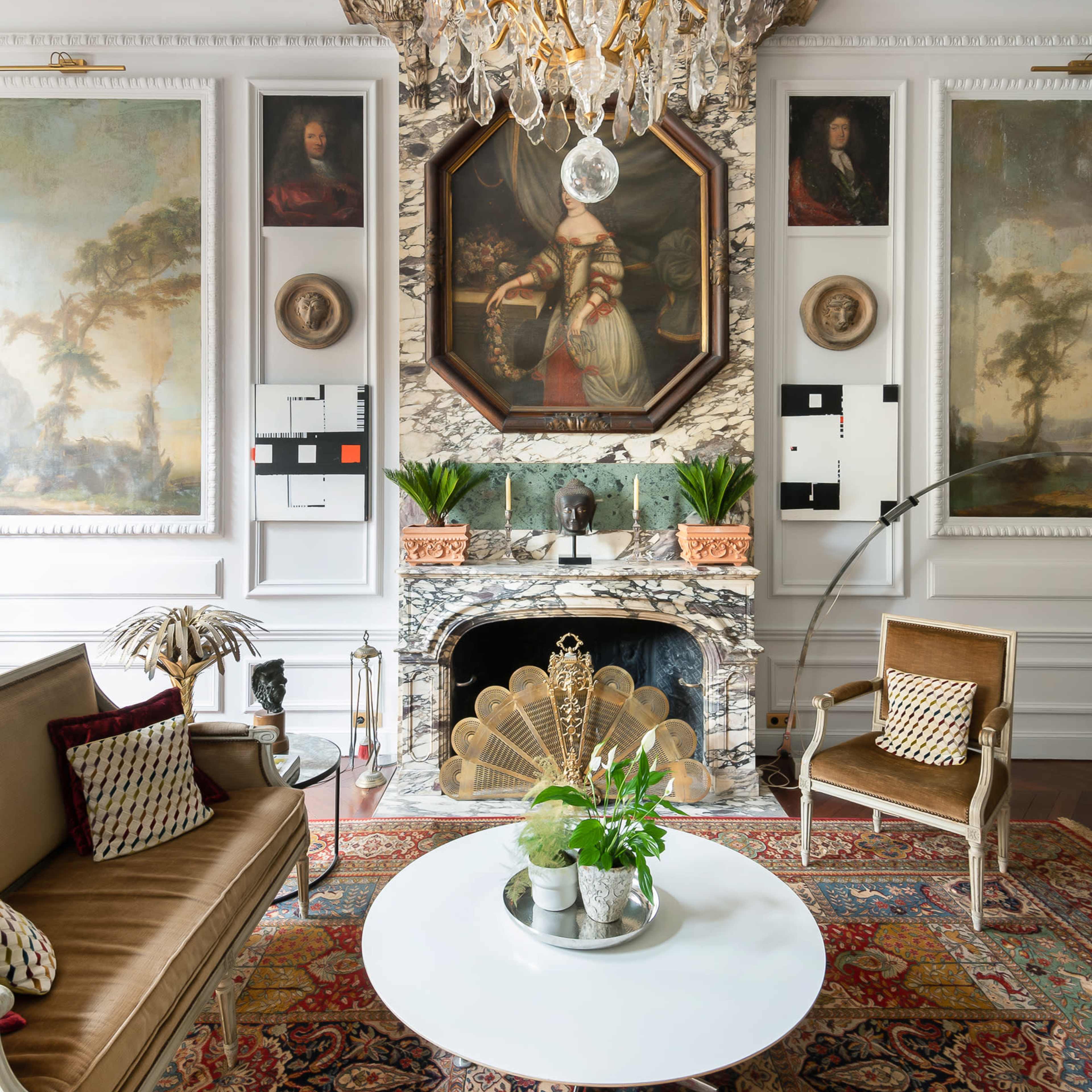 The living room features an ornate chandelier above a round white coffee table, surrounded by upholstered seating and framed artwork, with a decorative fireplace as the focal point.