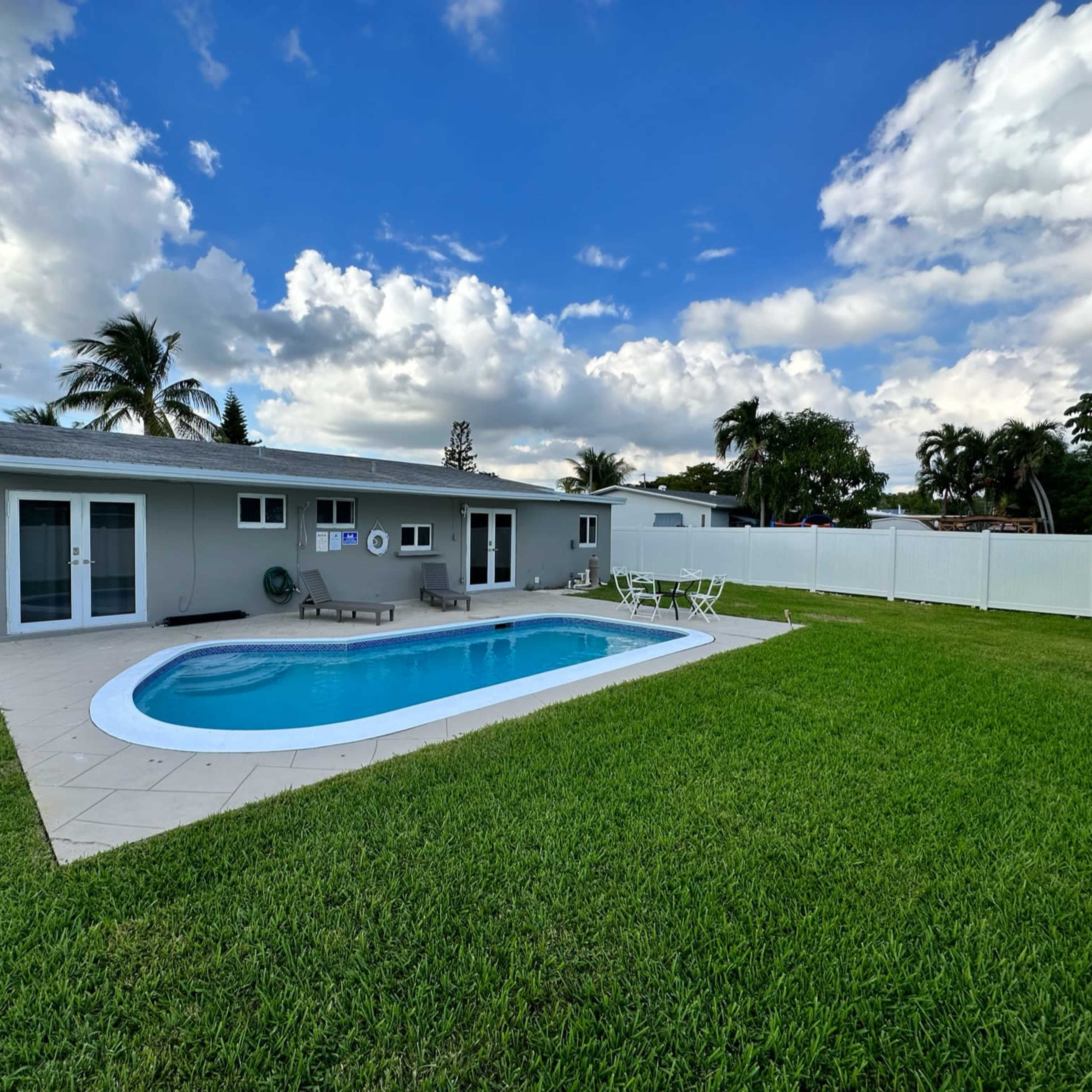 A single-story house with a pool surrounded by a green lawn and palm trees under a partly cloudy sky.