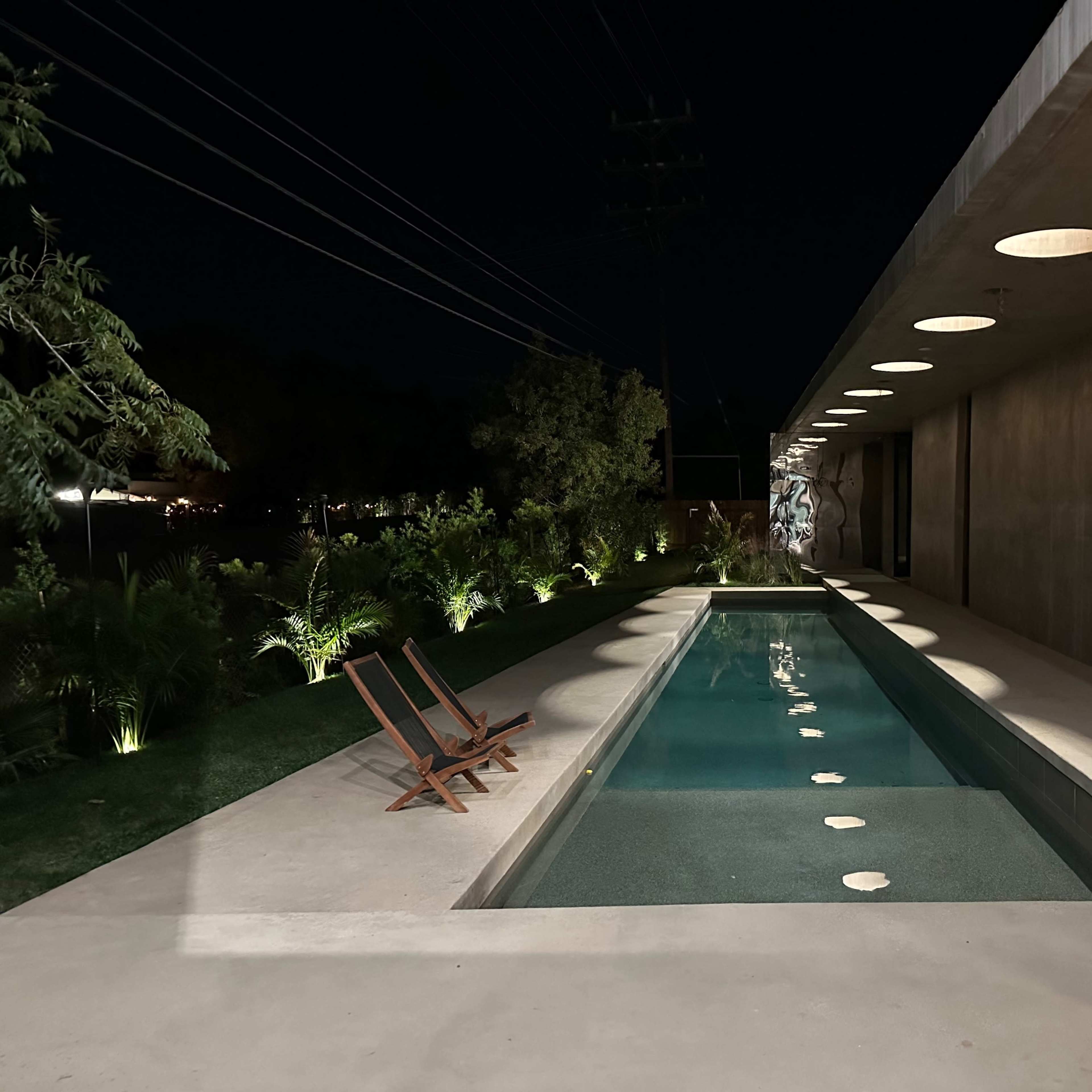 A modern, minimalist pool area is illuminated at night, featuring a rectangular pool surrounded by concrete and landscaped greenery.