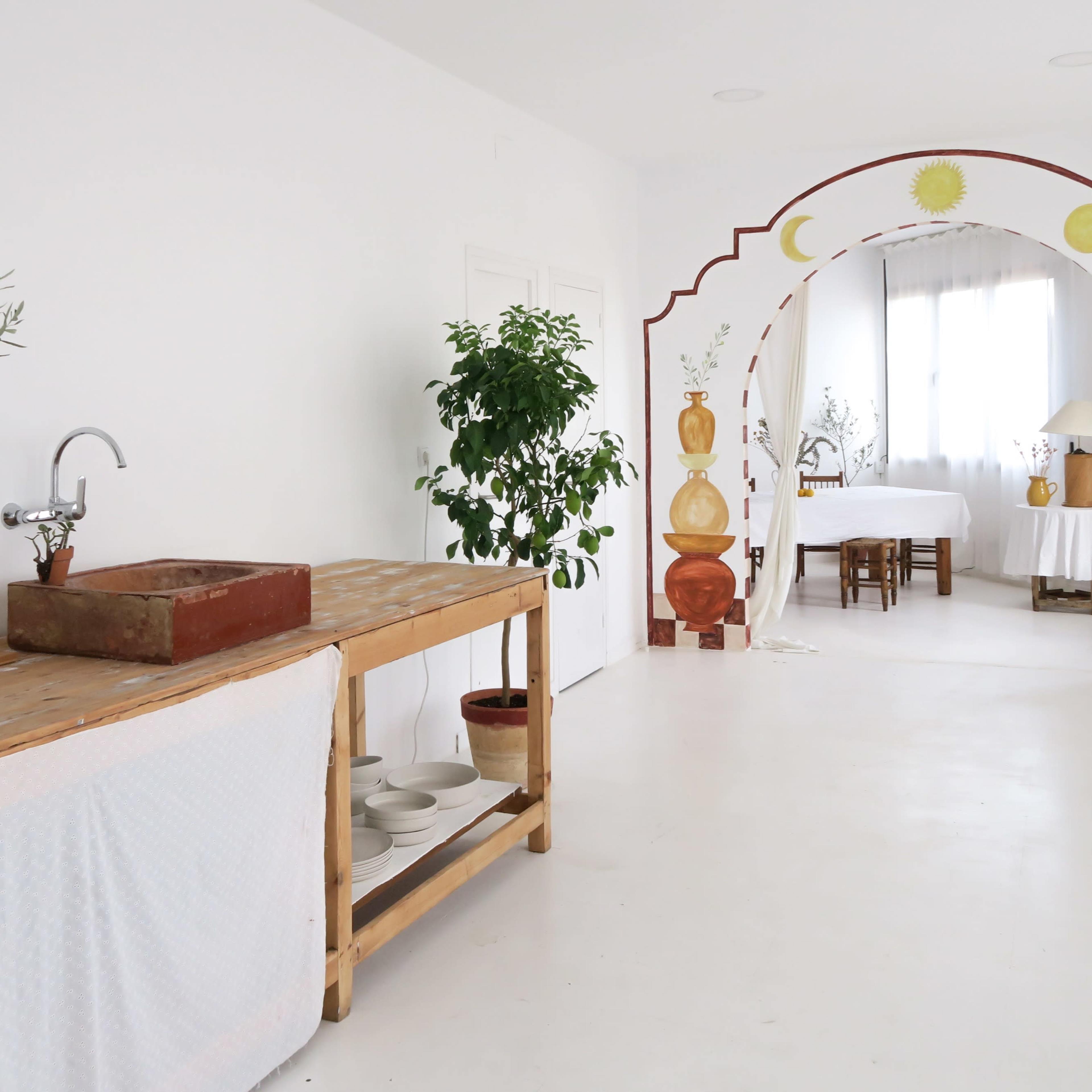 A spacious, airy room features a wooden table with a sink, potted plants, and an archway adorned with painted motifs leading to a dining area.