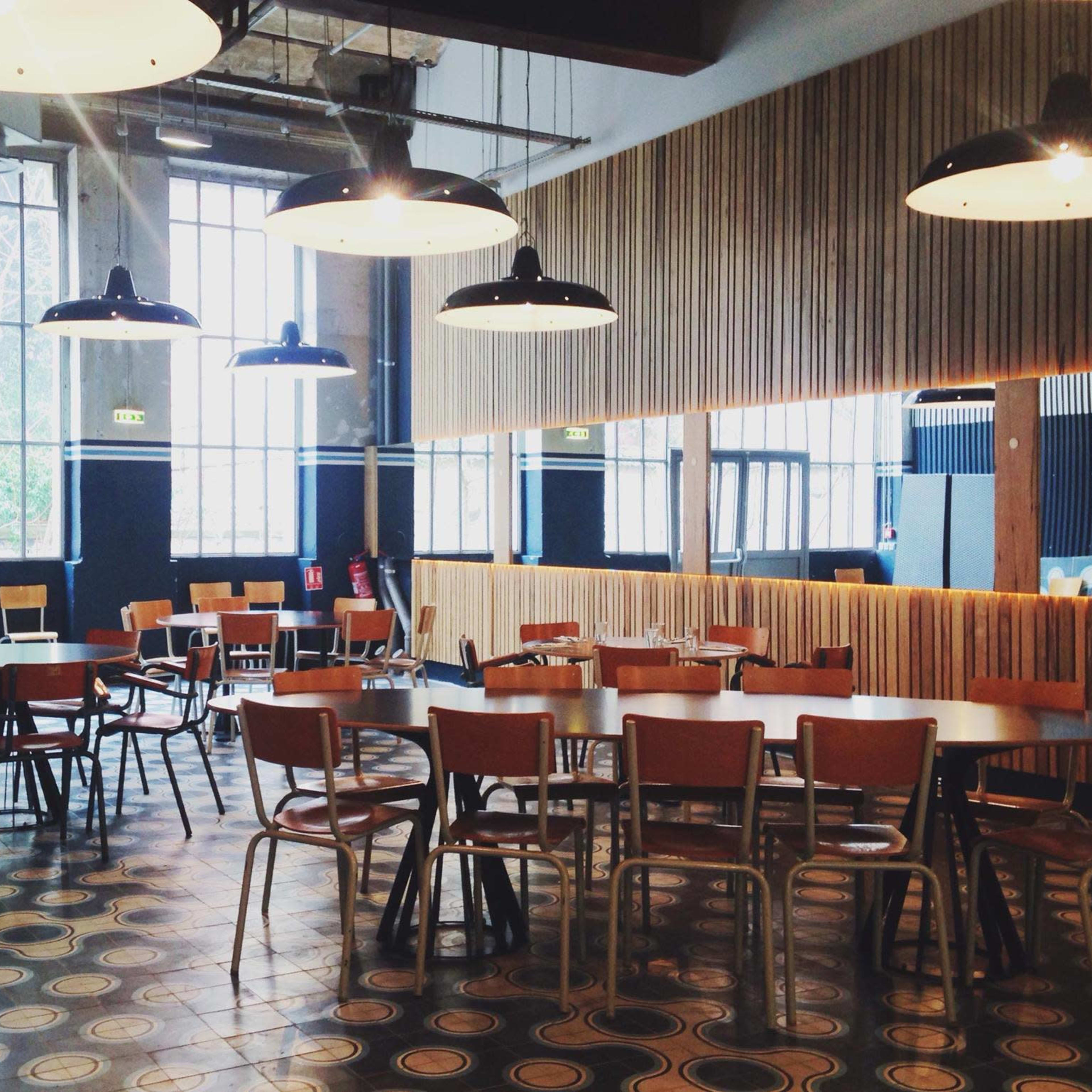 A spacious, modern café interior with several wooden tables and chairs, large windows allowing natural light, and industrial-style lighting fixtures.