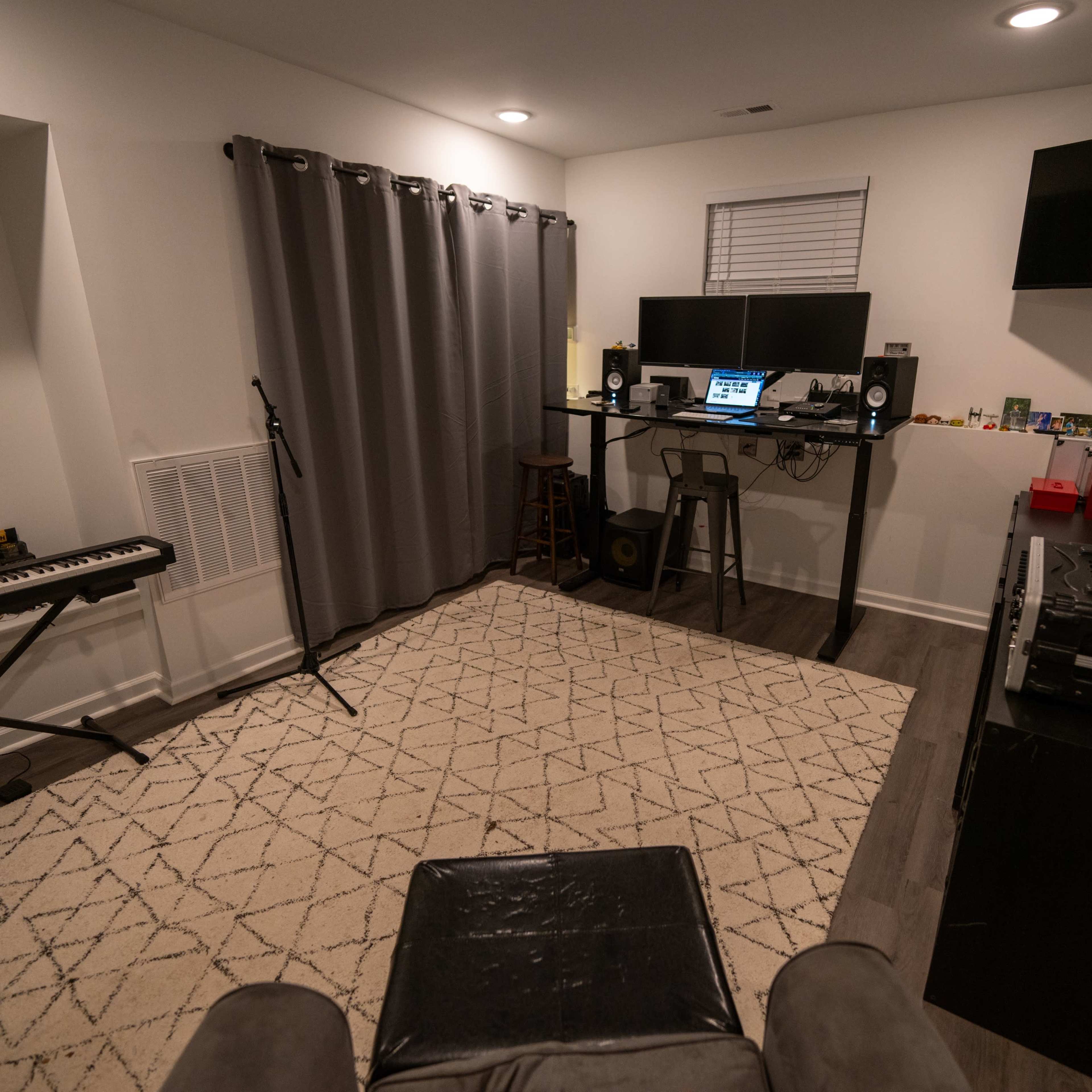 A home music studio with a piano, microphone, desk equipped with audio equipment, and a seating area, all set in a sparsely decorated room.