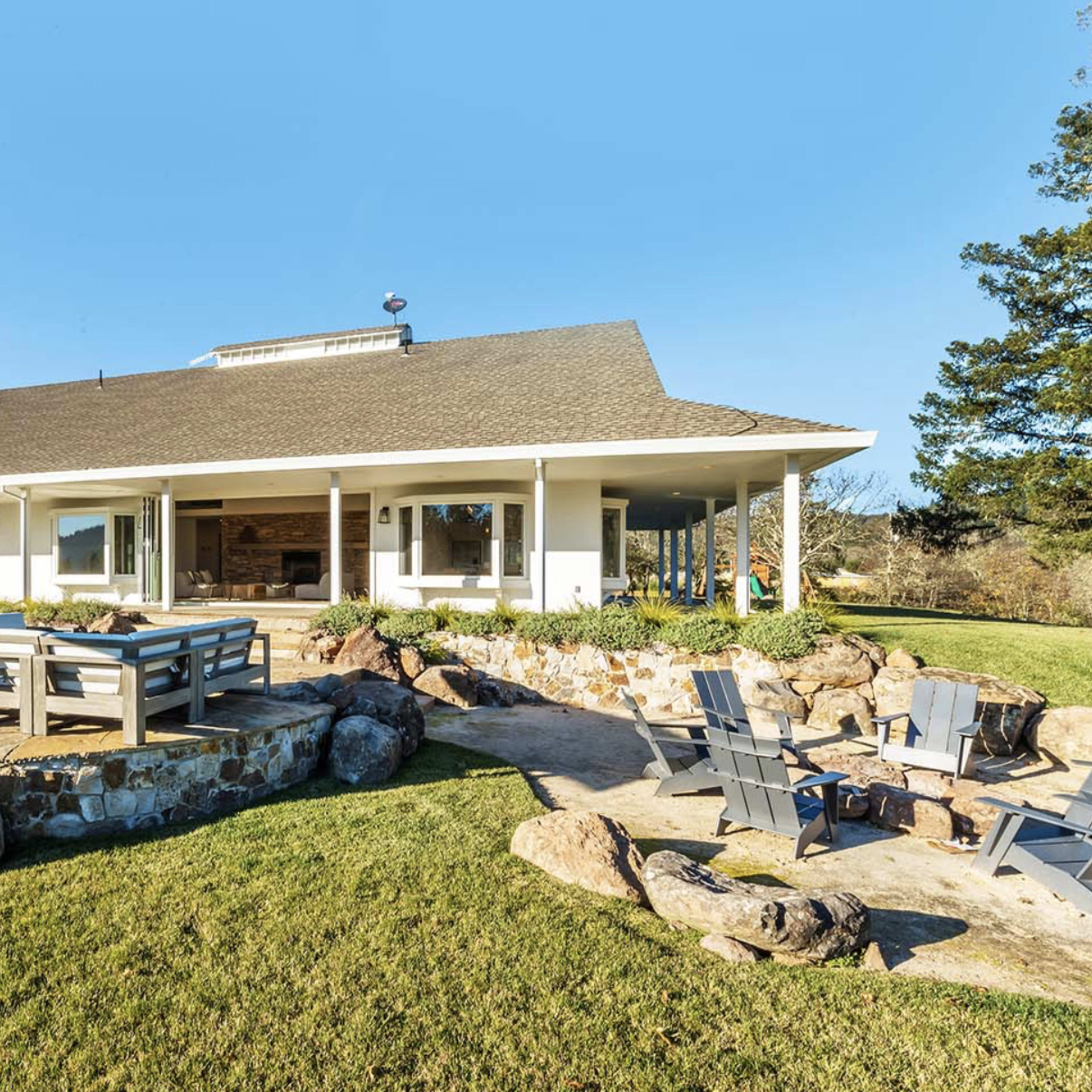 A single-story house with a spacious patio area featuring a dining table and stone landscaping is surrounded by a manicured lawn and trees.