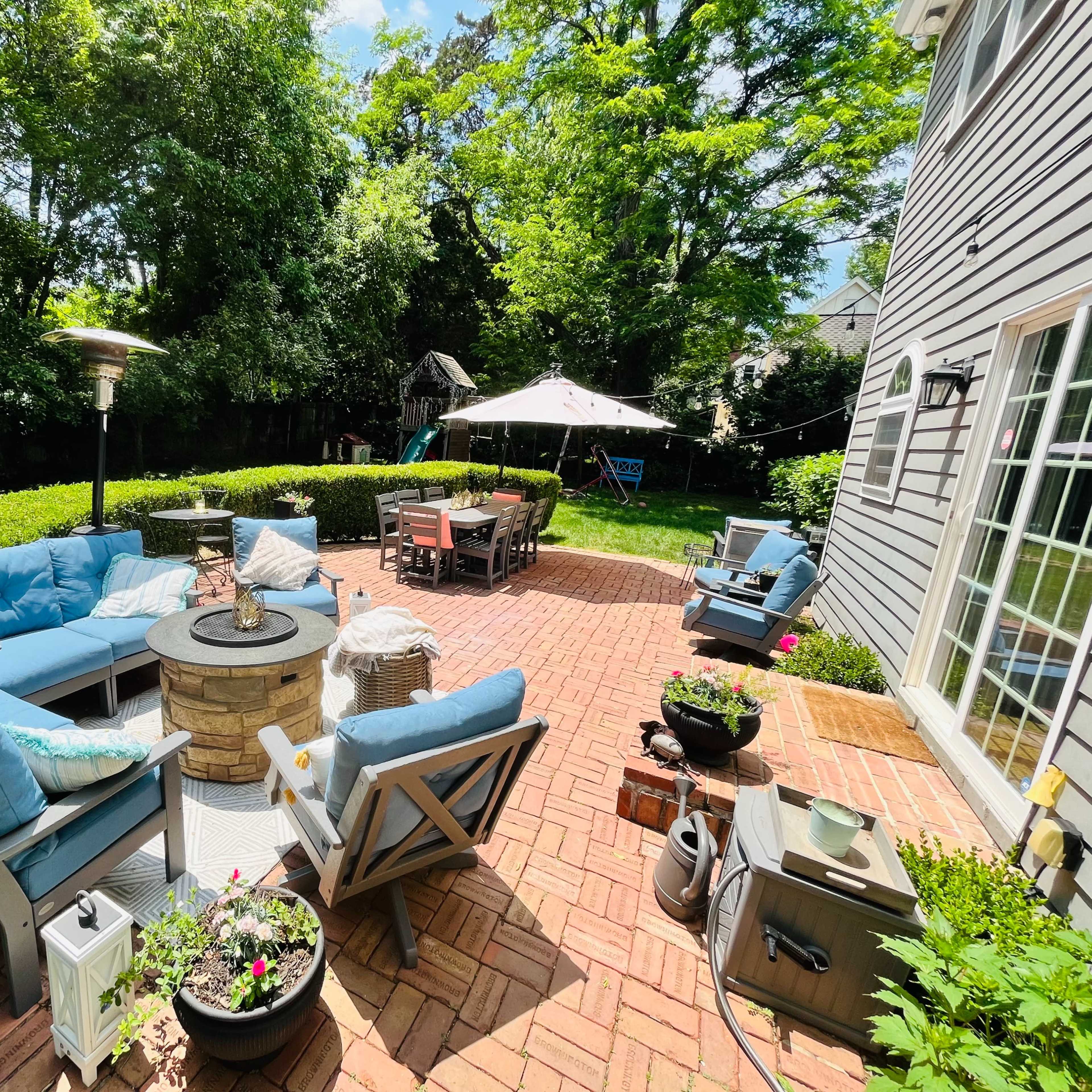 A backyard patio features a blue sectional sofa arranged around a stone fire pit, surrounded by greenery and outdoor dining furniture under a canopy.