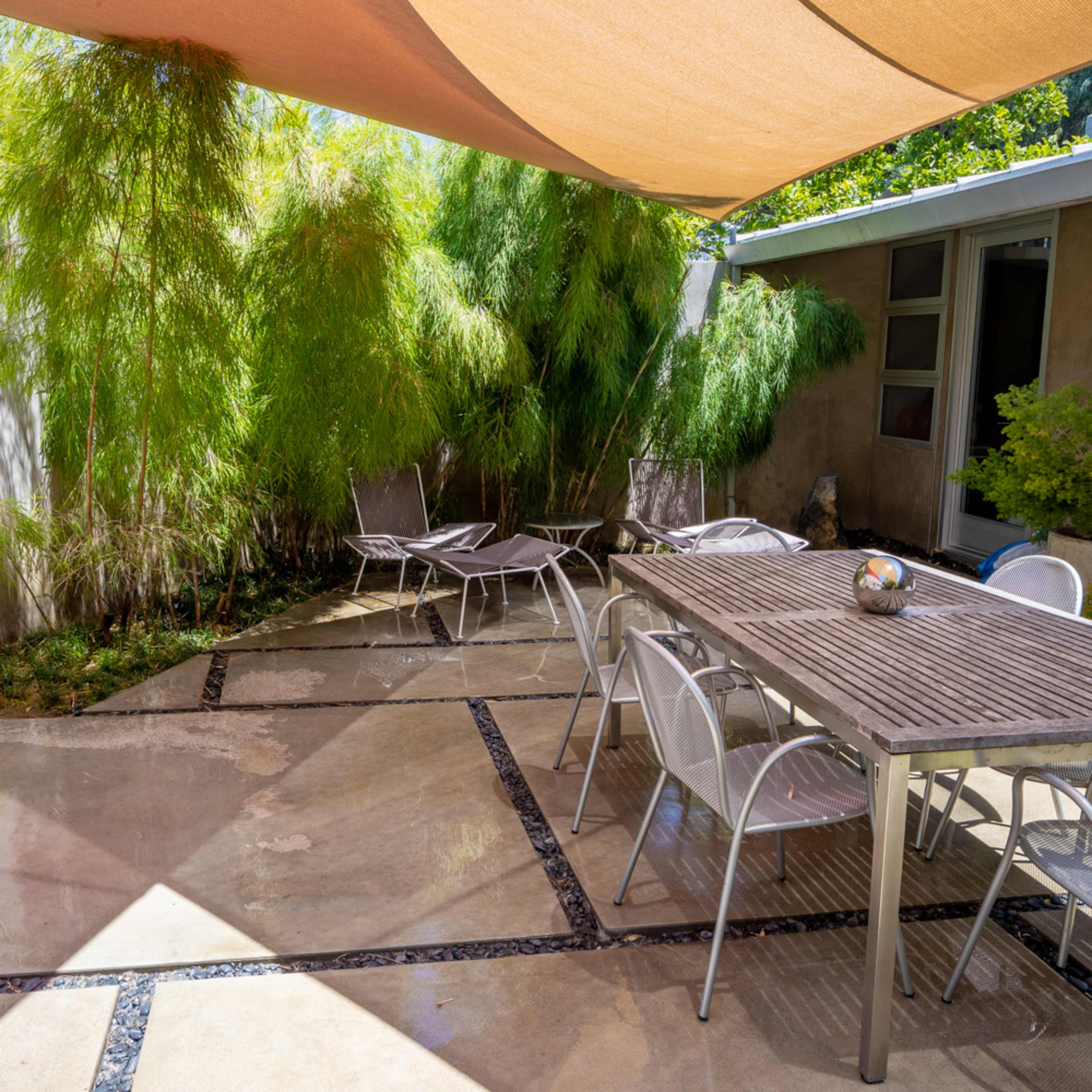 A patio area features a table surrounded by chairs under a shade sail, with bamboo plants and concrete flooring.