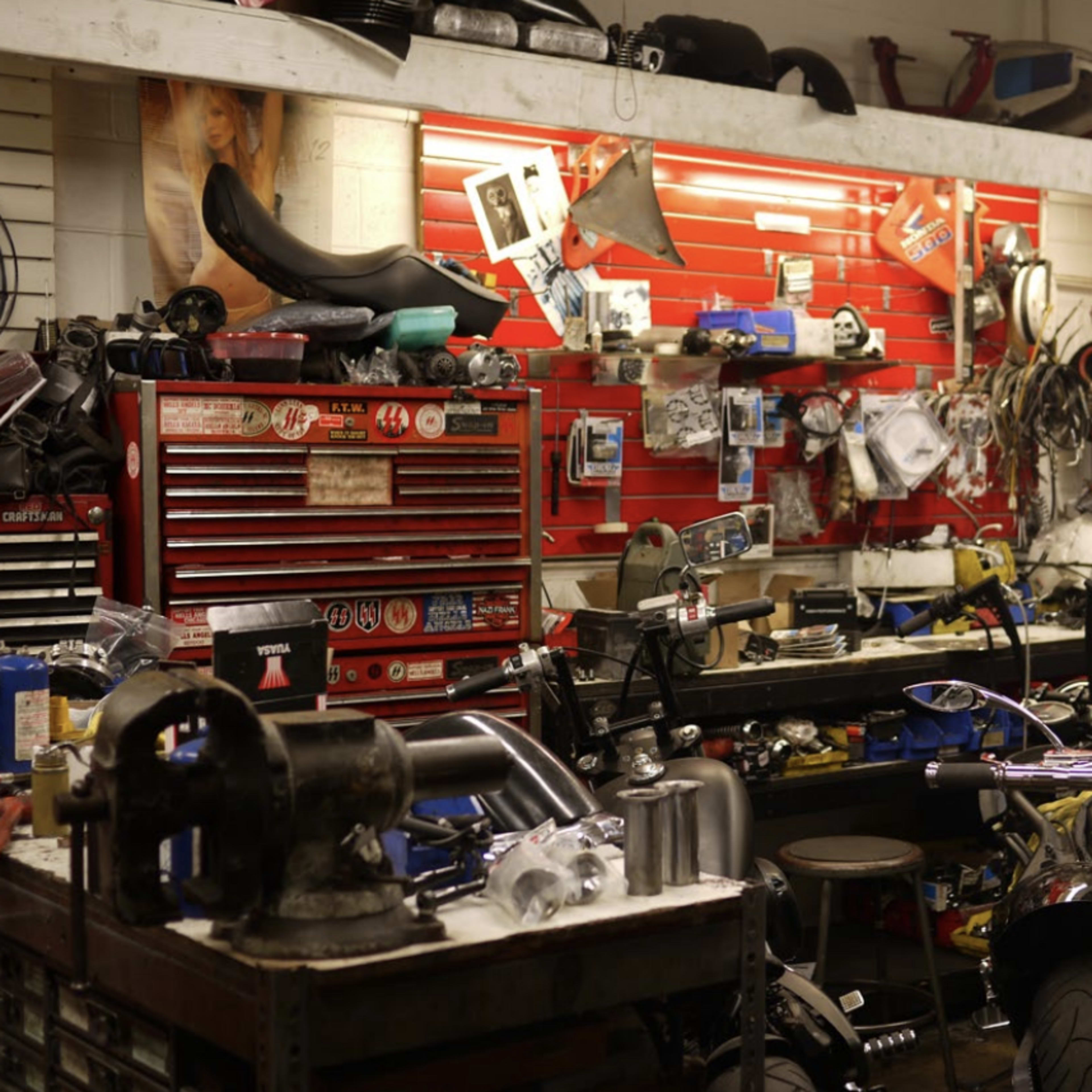 A cluttered motorcycle repair shop features various tools, parts, and workbenches, with a red tool chest prominently displayed against the wall.