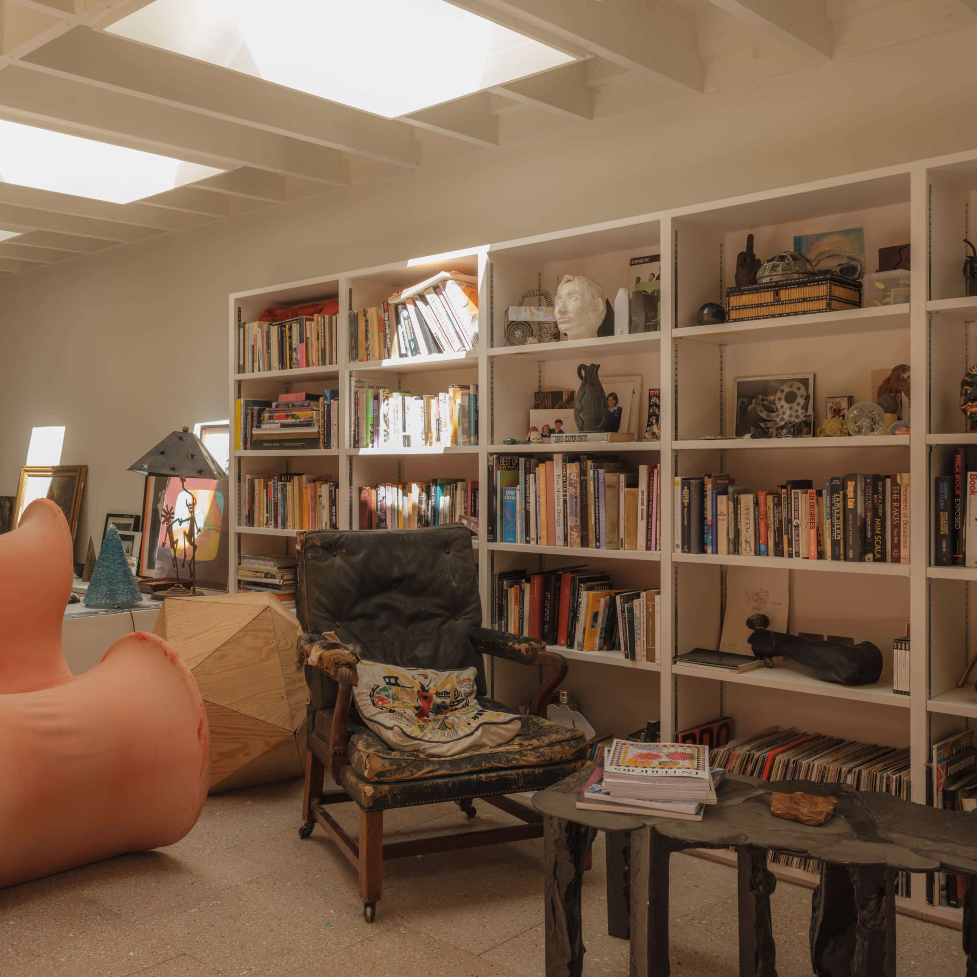 A modern living room featuring a curved pink chair, a black leather armchair, a wooden table, and shelves filled with books and decorative items.