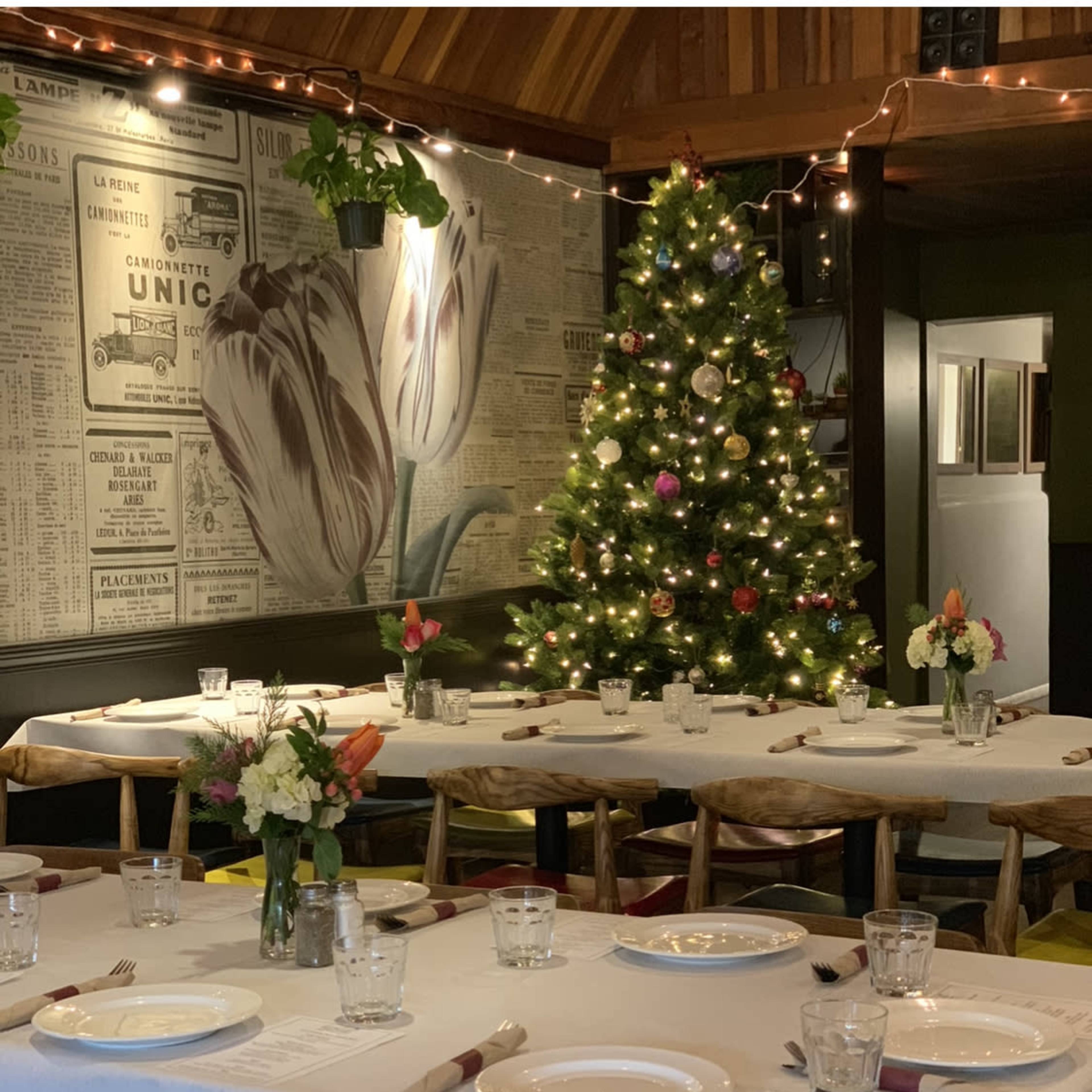 A dining area features neatly arranged tables with white tablecloths, floral centerpieces, and a decorated Christmas tree in the background.