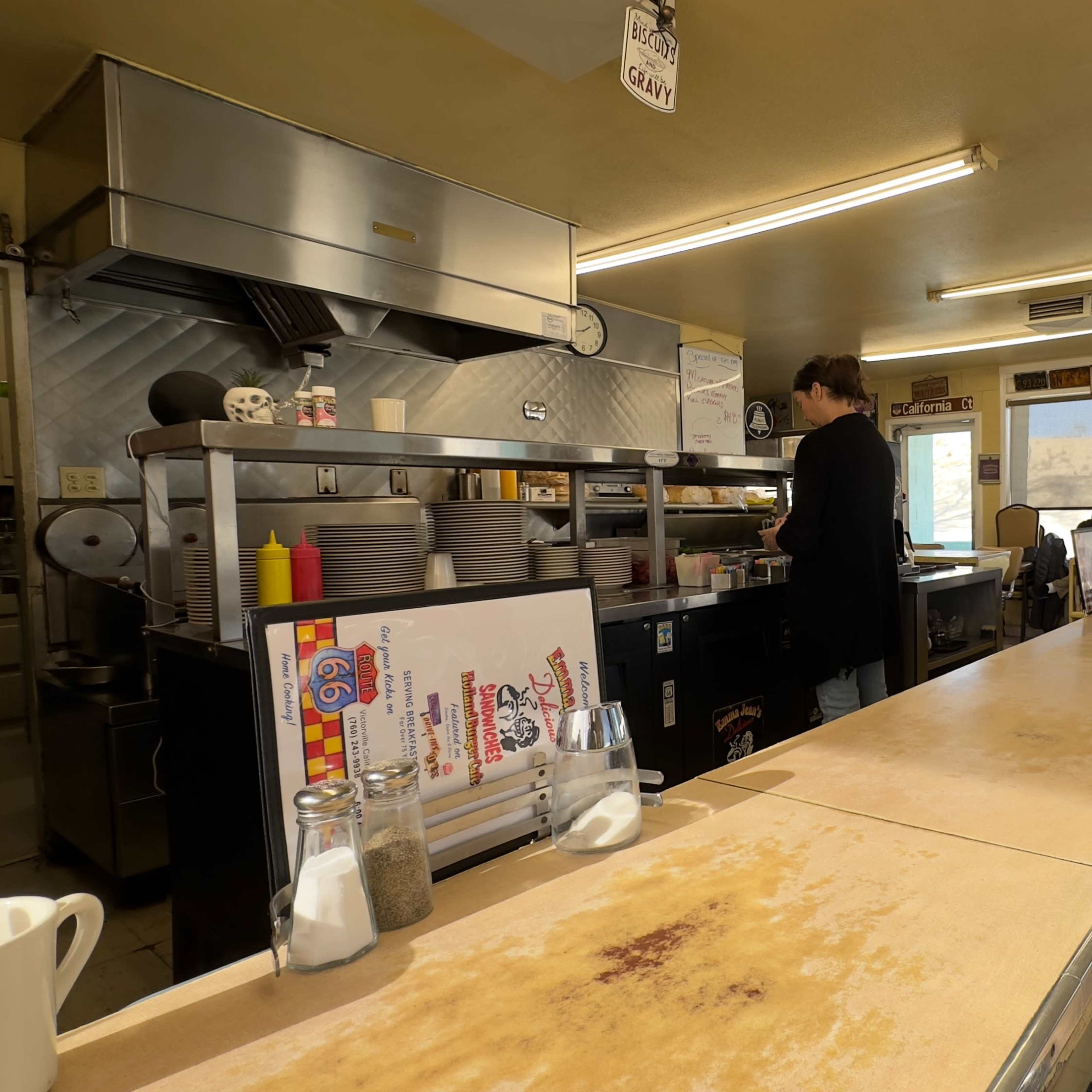 The interior of a diner with a long counter, kitchen area in the back, and several patrons seated by large windows.