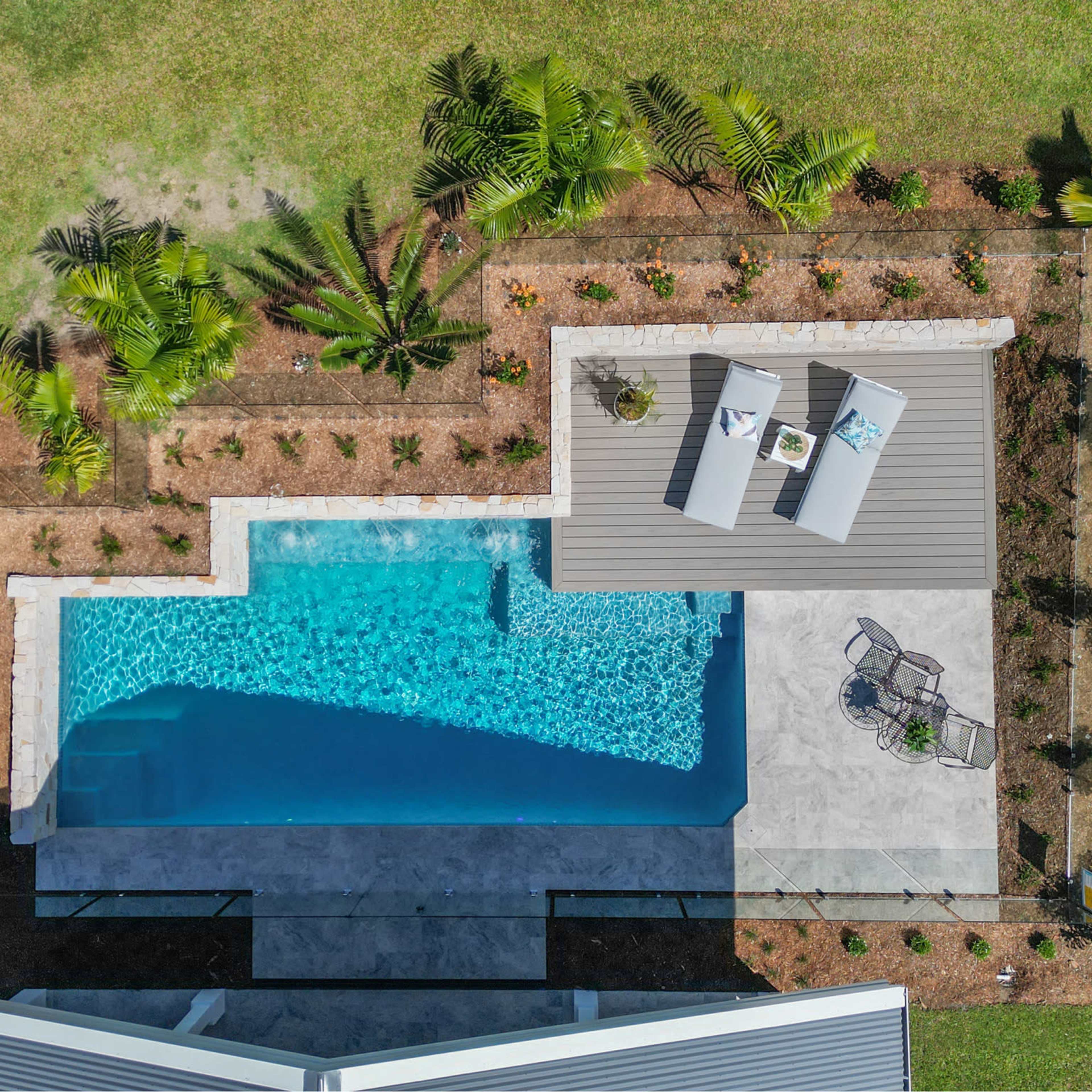 An aerial view shows a rectangular swimming pool surrounded by a landscaped area with palm trees and lounge chairs.