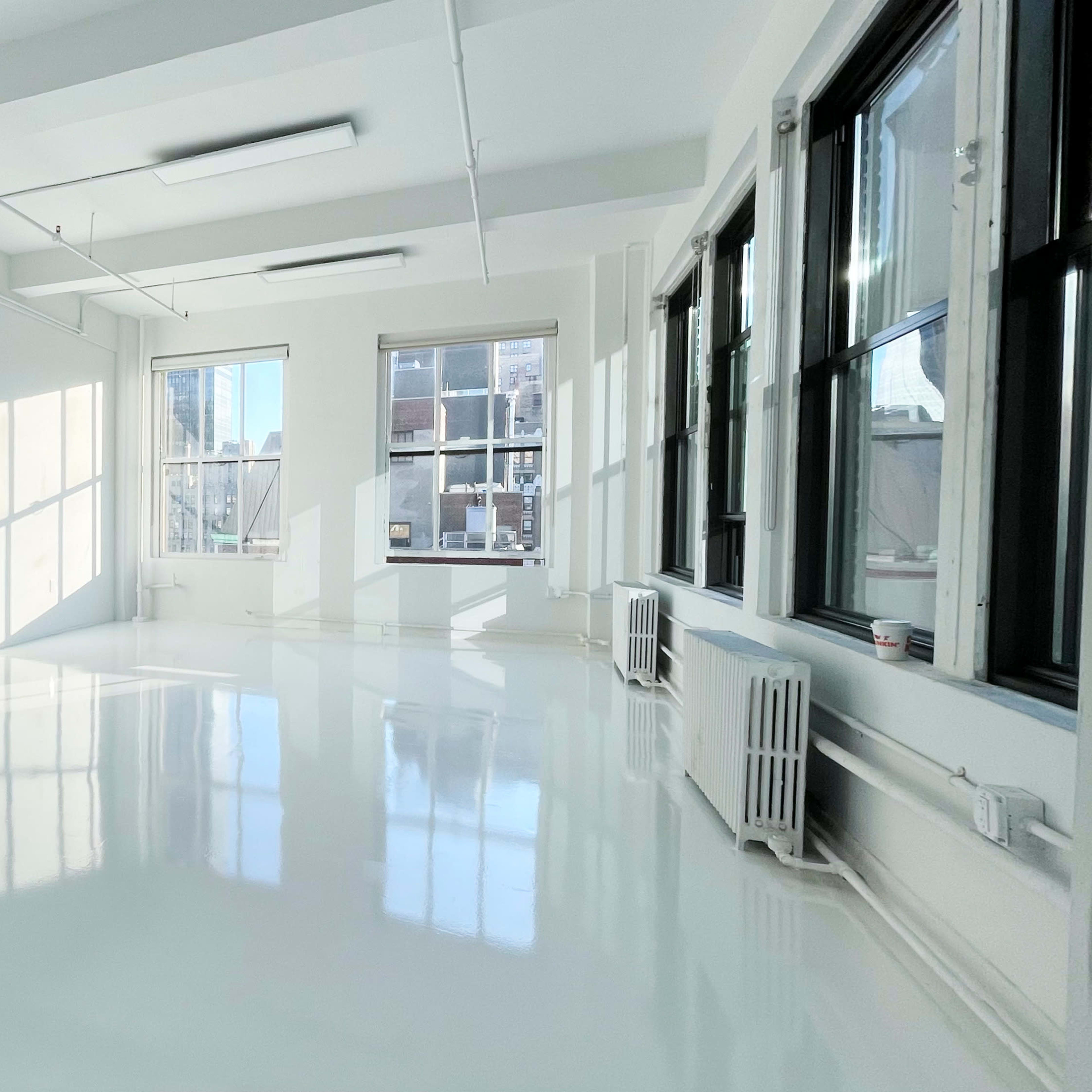 A spacious, well-lit room with white walls and a glossy white floor, featuring large windows that allow sunlight to enter.