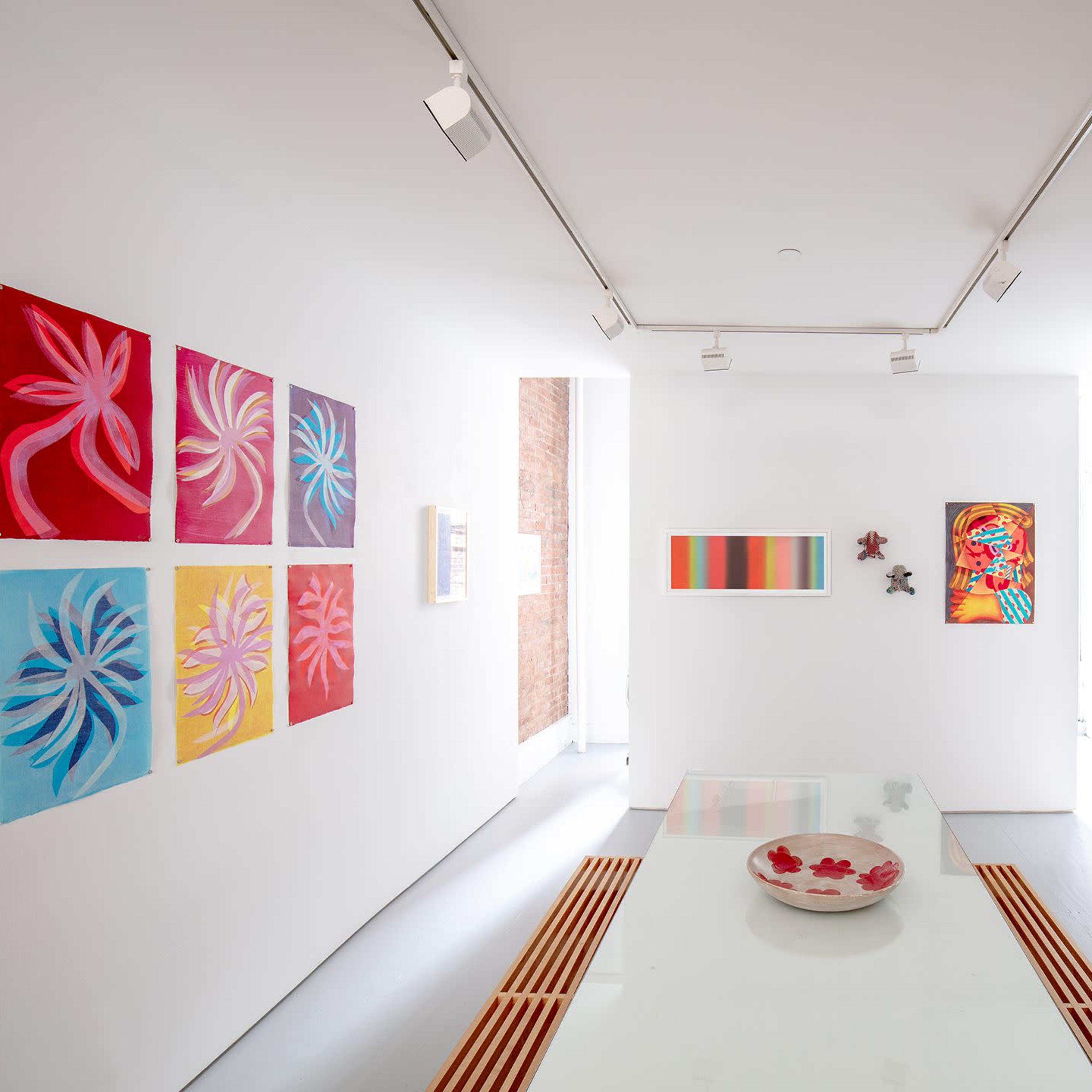 A brightly lit gallery space featuring colorful paintings on the walls and a glass table with a decorative bowl in the center.