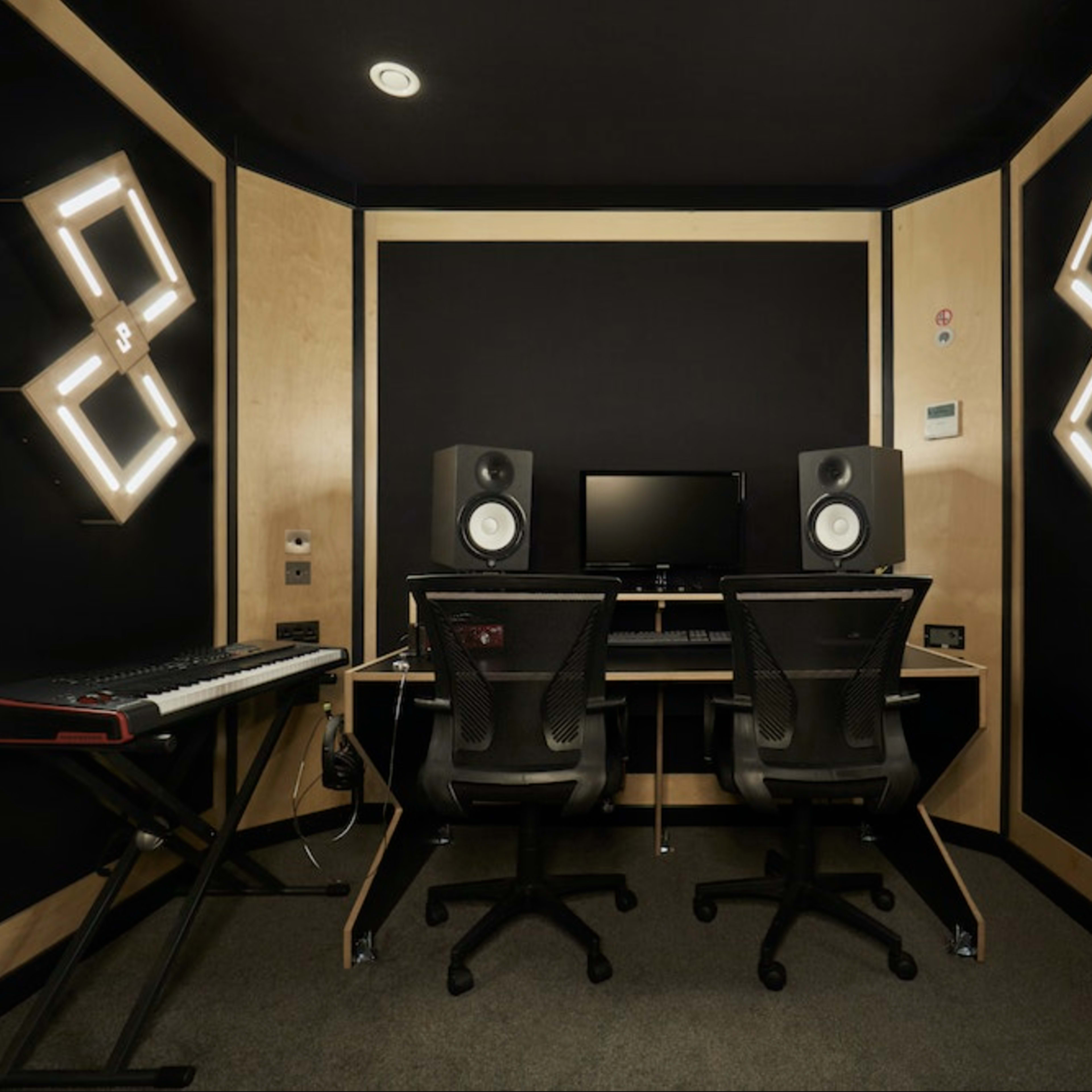 A music production studio with two black chairs, a computer monitor, speakers, and a keyboard in a small, dark room.