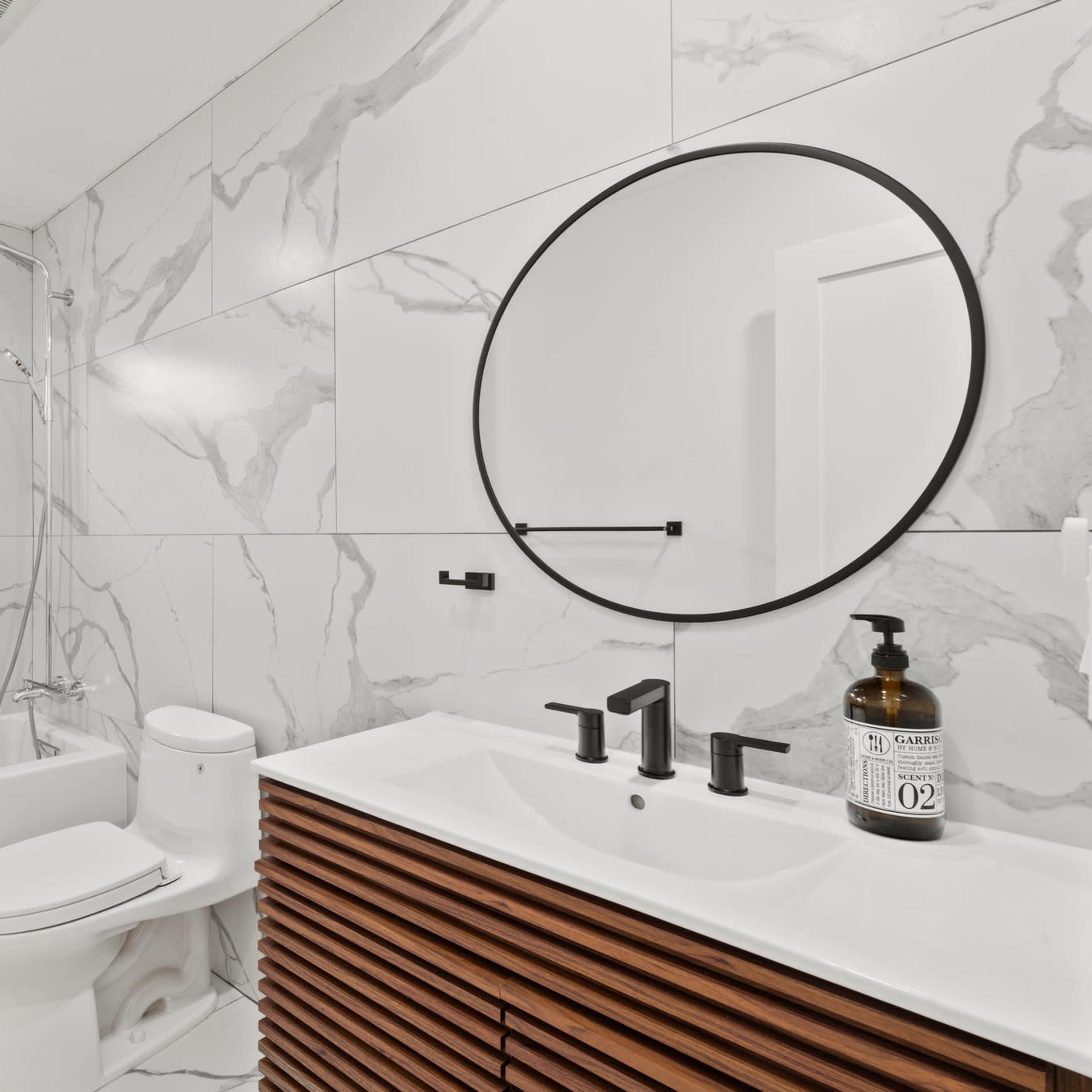A modern bathroom featuring a freestanding bathtub, a circular mirror, a wooden vanity with black faucets, and marble-patterned walls.