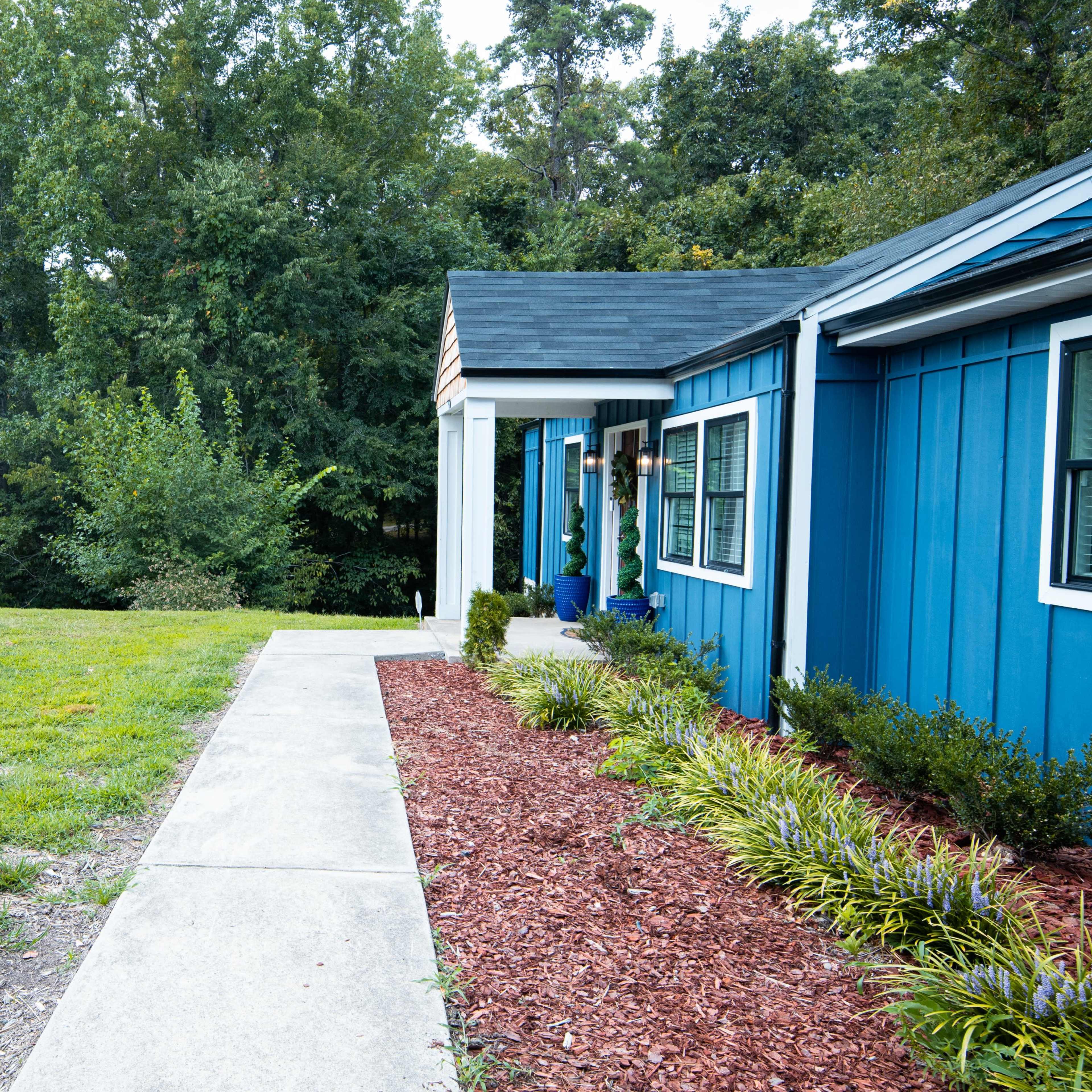 A blue house with a walkway leads to a grassy area and trees in the background.