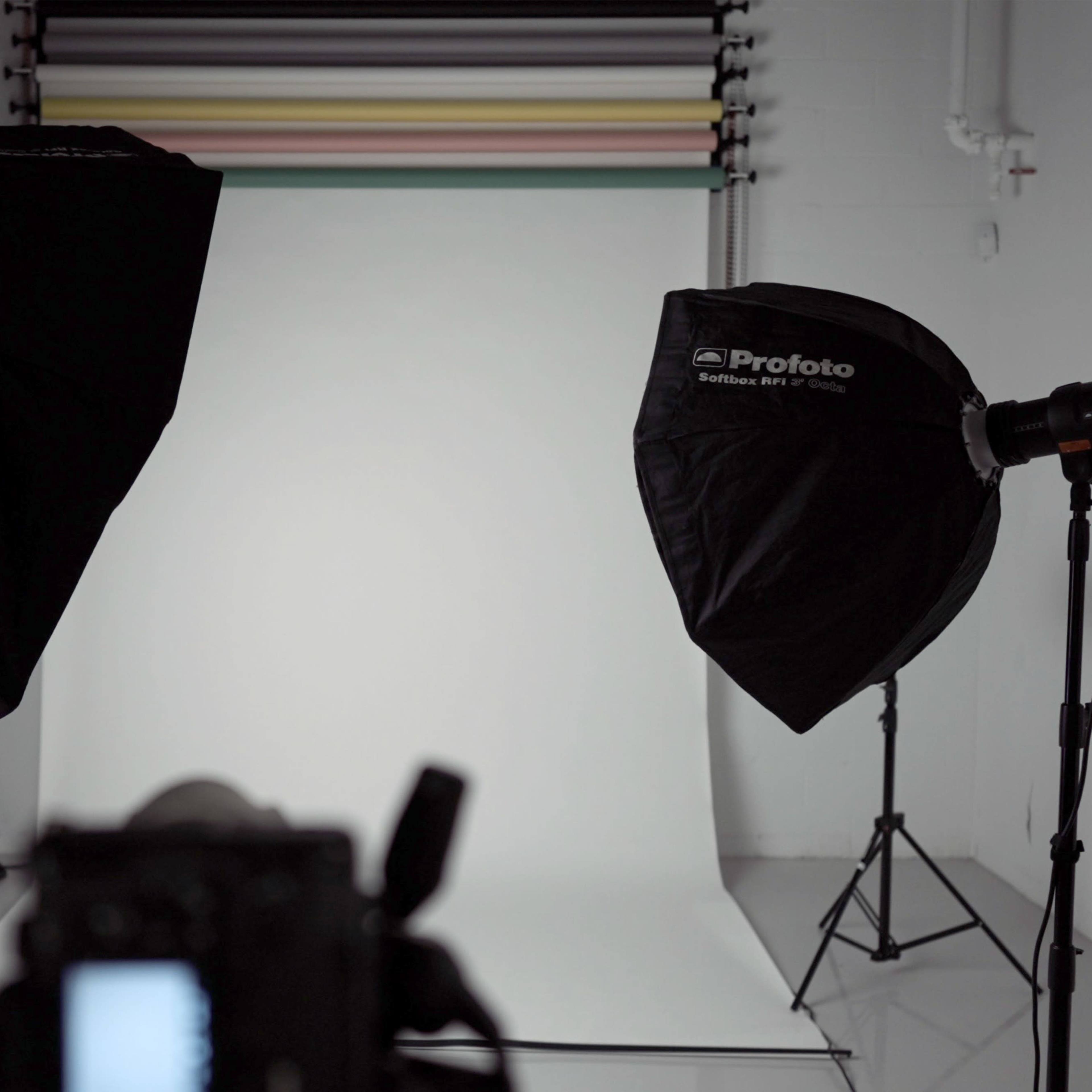 A photography studio setup features two softbox lights and a camera positioned in front of a neutral backdrop.