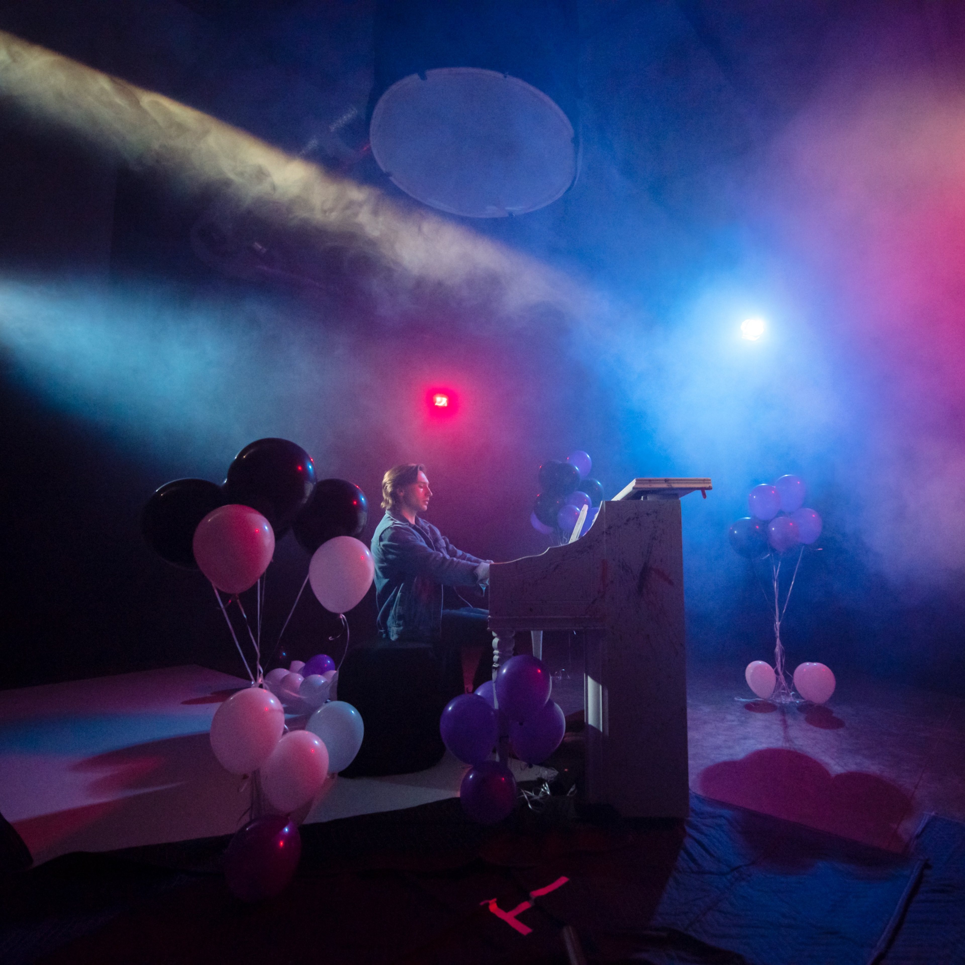 A musician plays a piano on a stage decorated with black, white, and purple balloons, surrounded by colored lighting and fog.