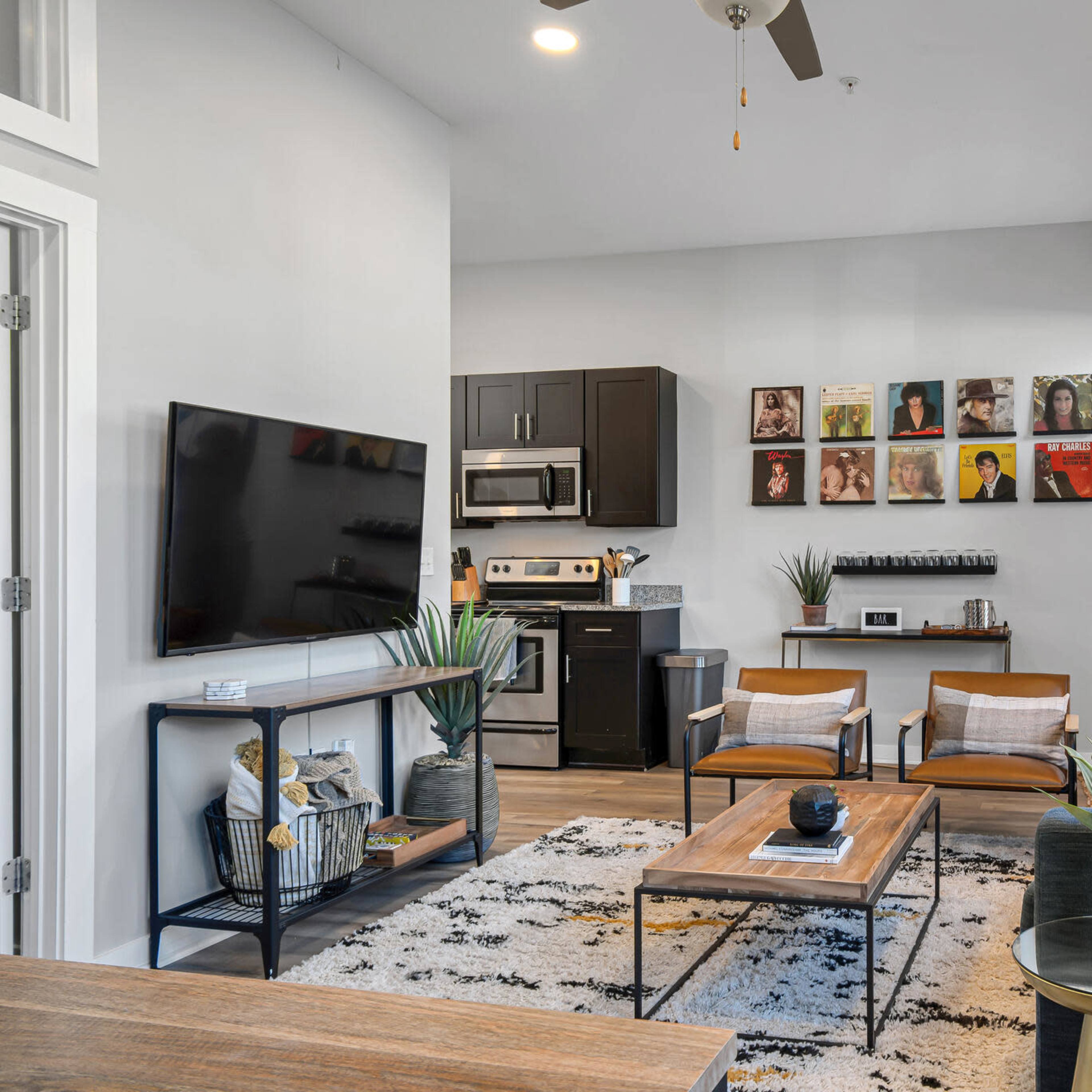 The living room features a modern design with a television, a kitchen area, two armchairs, and a coffee table, surrounded by decorative plants.