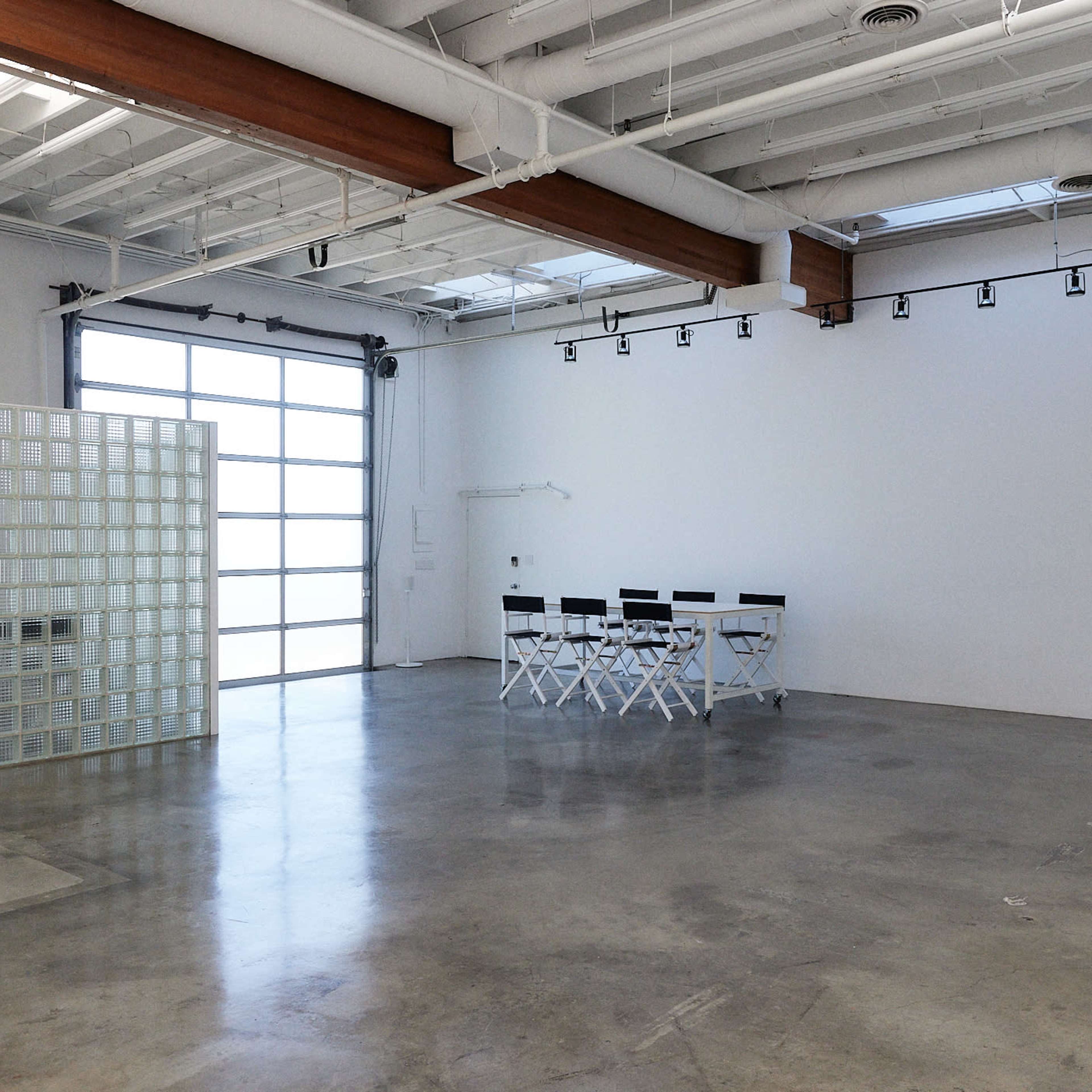 A spacious, modern room with concrete flooring, a glass block partition, and a row of white folding chairs against a wall of large windows.