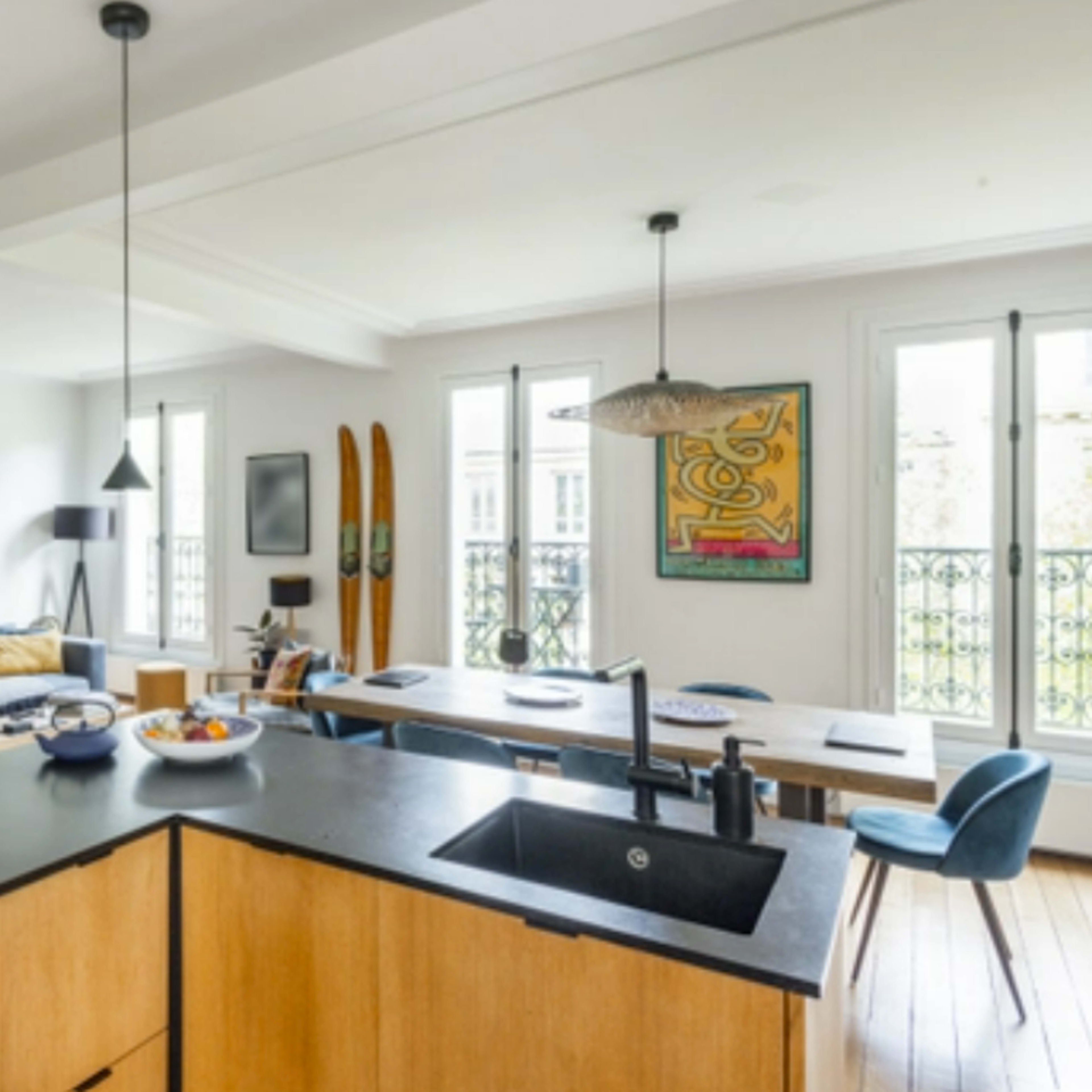 A modern kitchen with wooden cabinetry and a black countertop, connected to a bright, open living space featuring a sofa, artwork, and a dining area with skis adorning the wall.