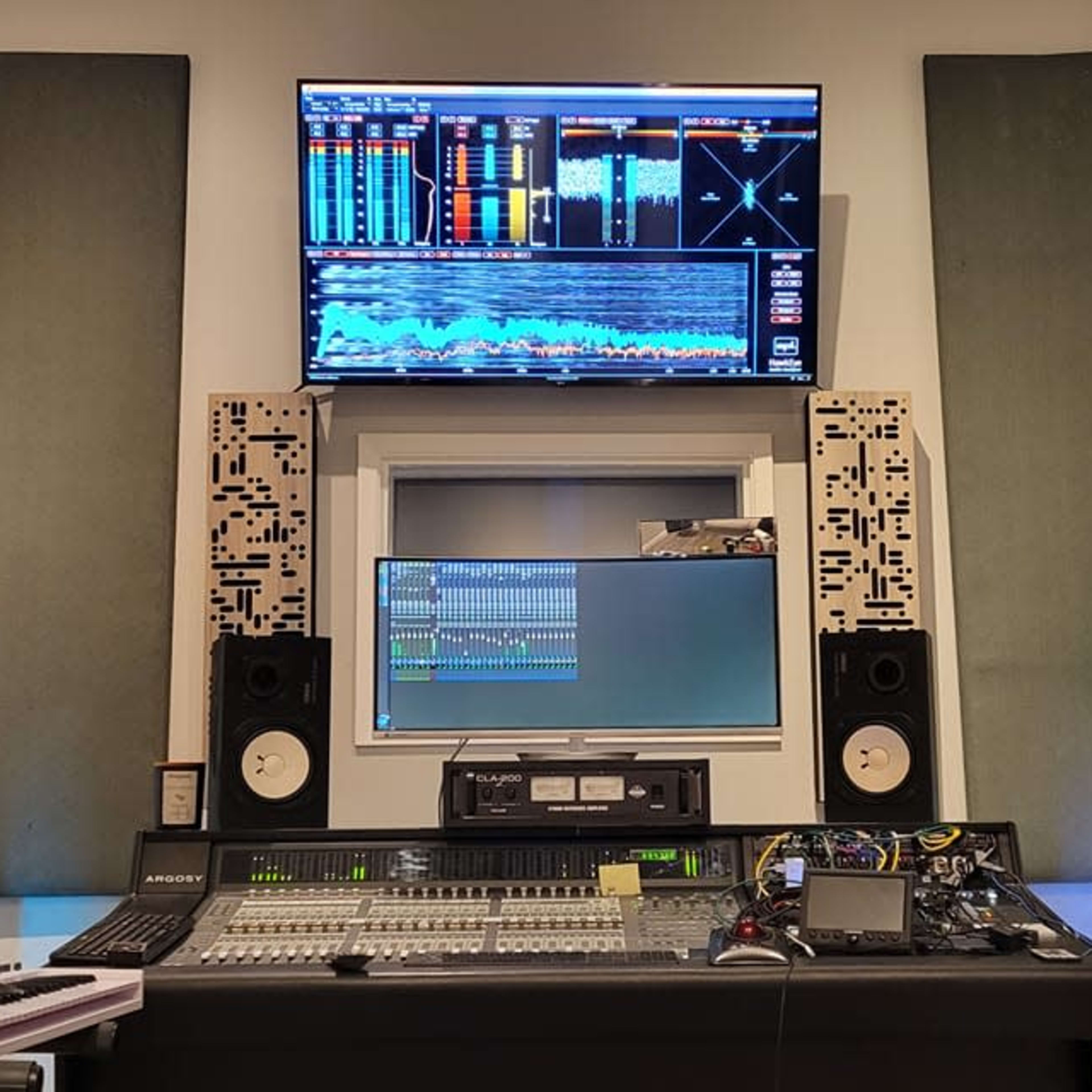 A music studio with a sound mixing console, computer monitors, and soundproofing panels on the walls.