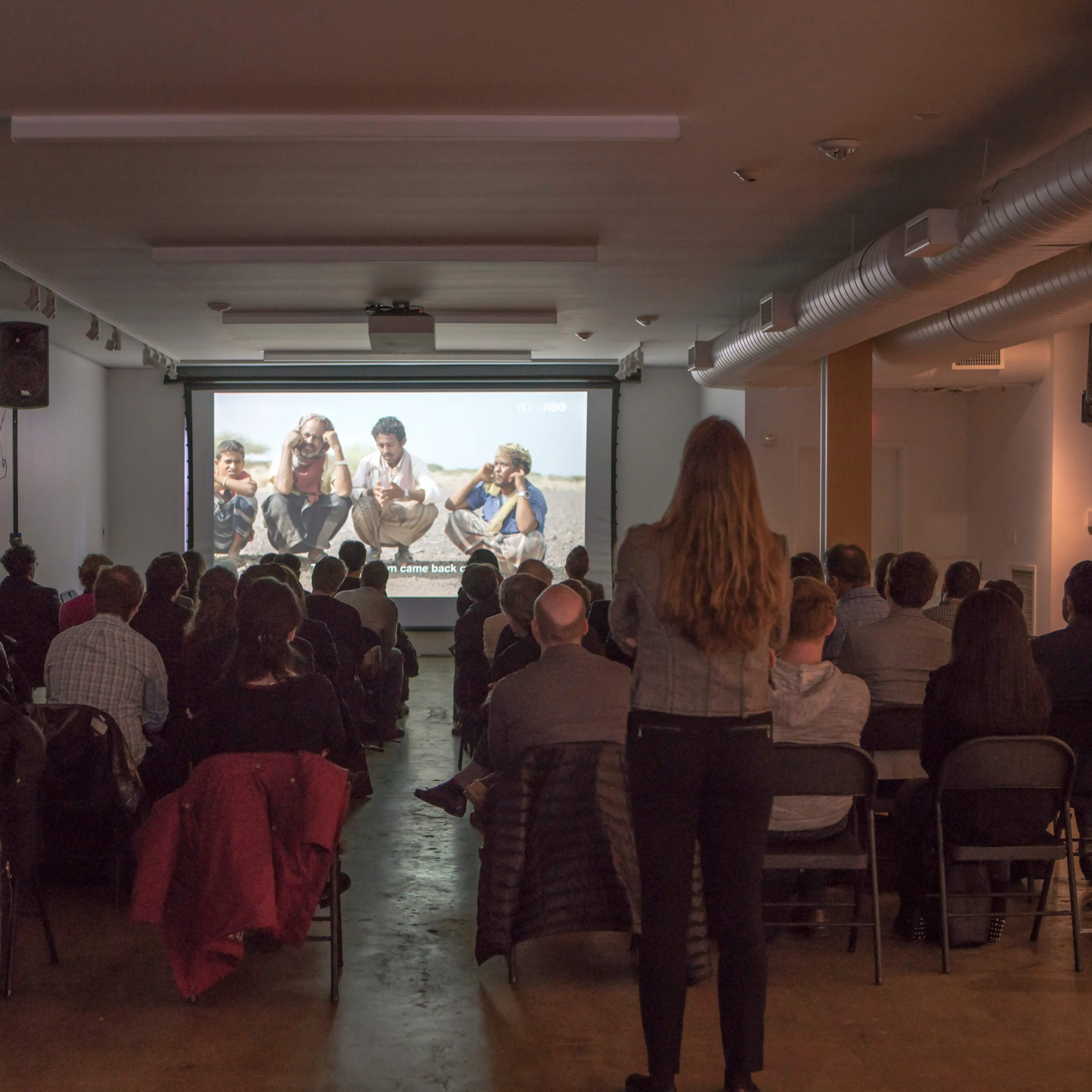 A crowd of people watches a presentation on a large screen in a well-lit indoor space.