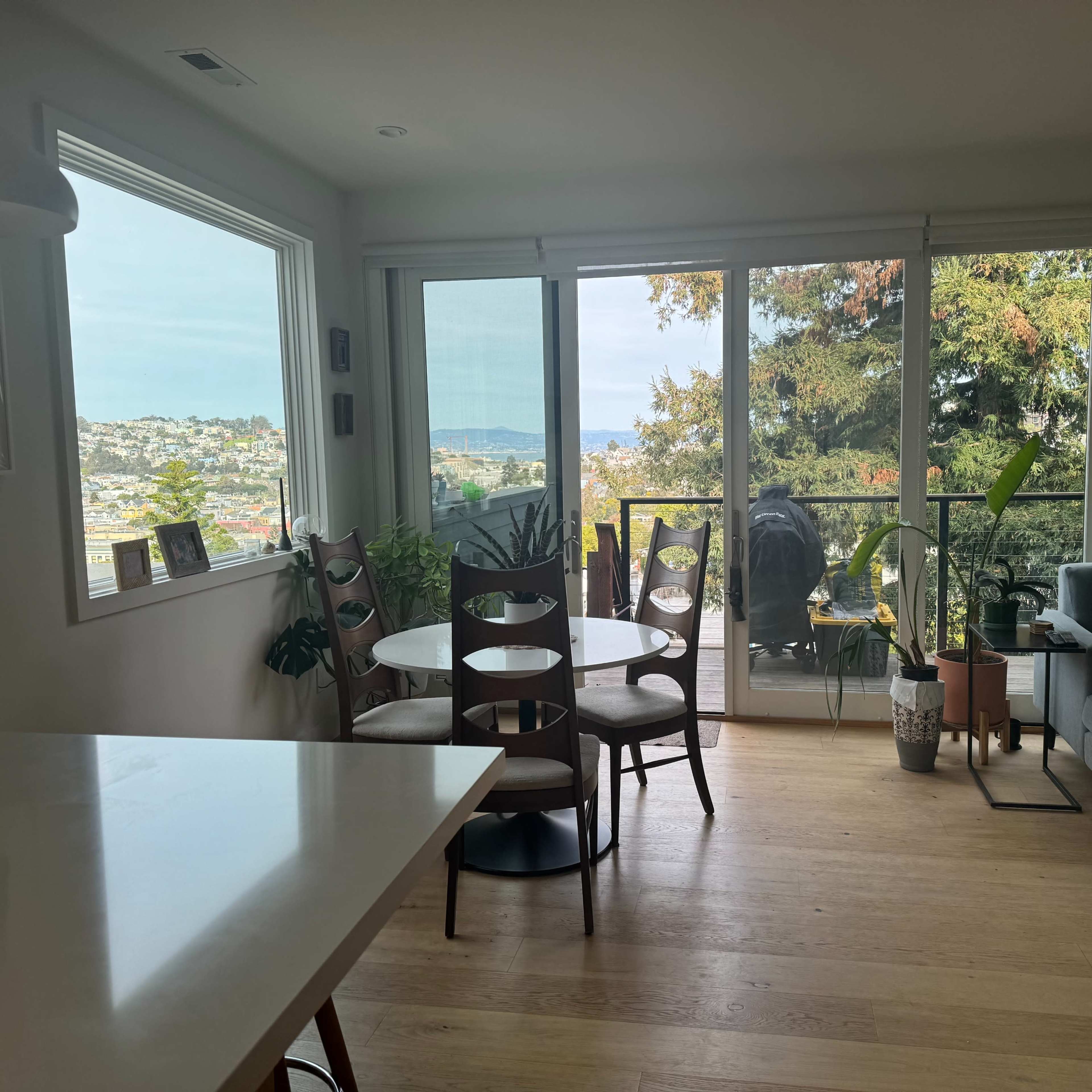 A bright living space featuring a dining area with wooden chairs, a modern couch, and large windows overlooking a scenic view with trees and water in the distance.