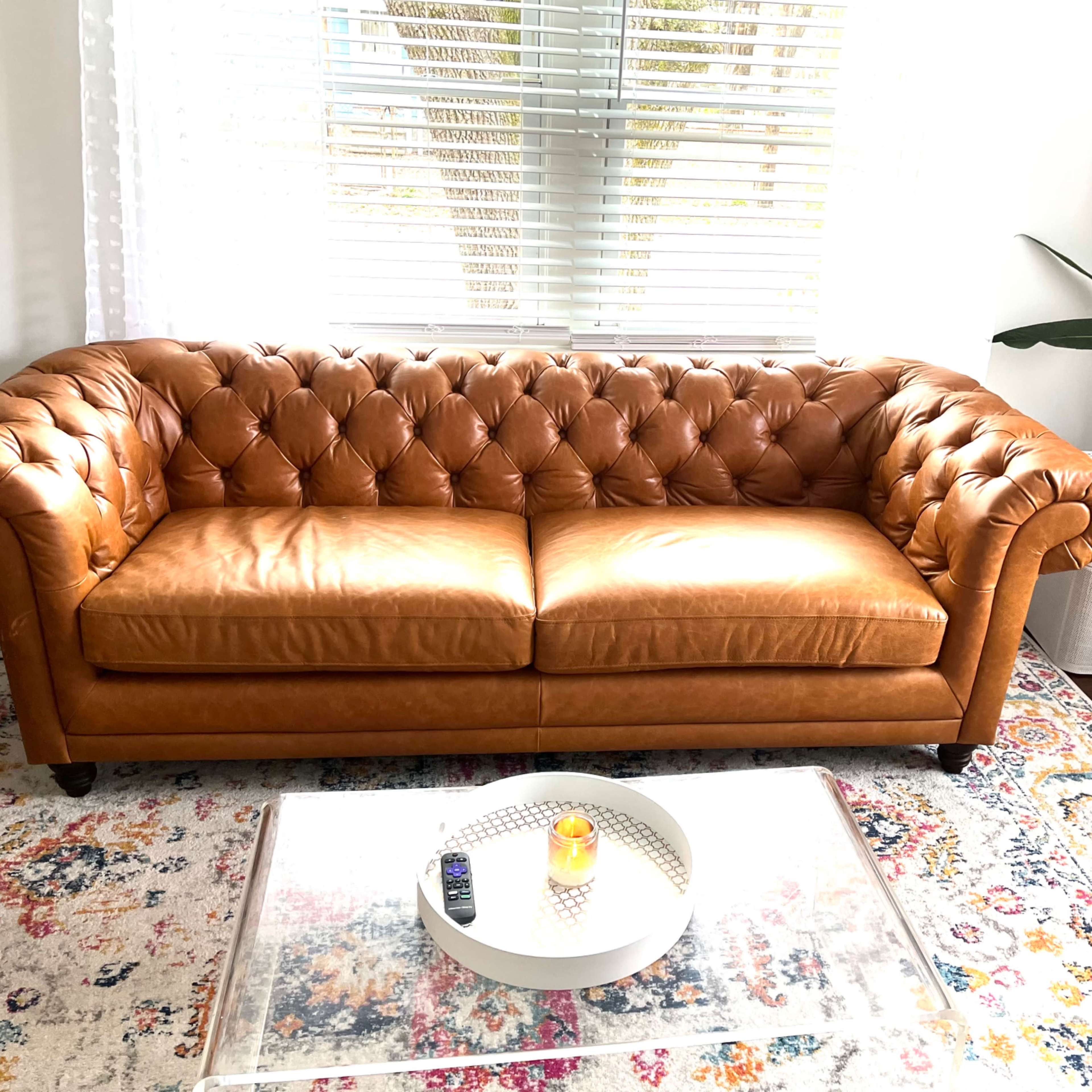 A tufted leather sofa sits in a well-lit room with a glass coffee table, a decorative candle, and a potted plant nearby.
