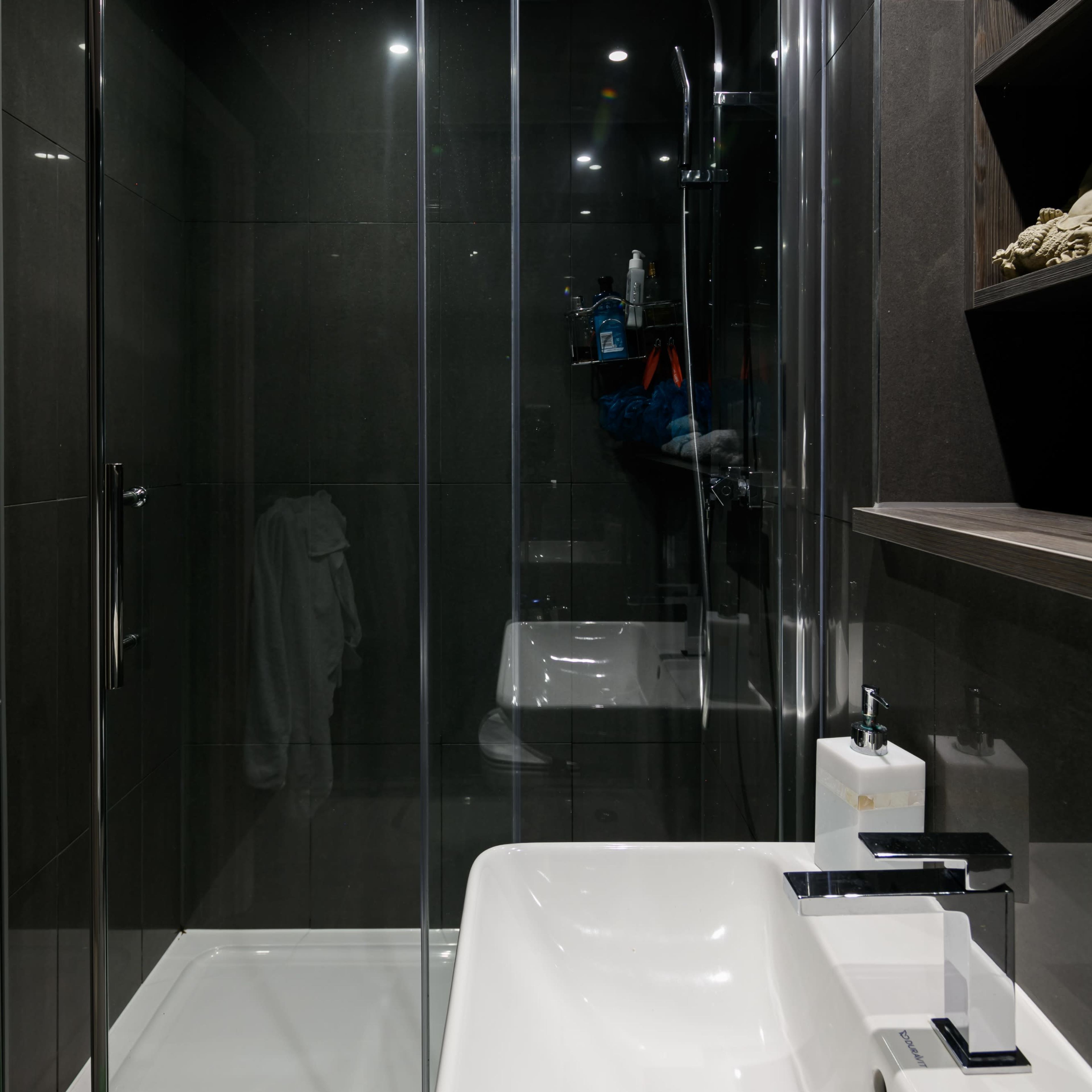 A modern bathroom features a glass shower enclosure, a white sink with contemporary fixtures, and dark tiled walls.