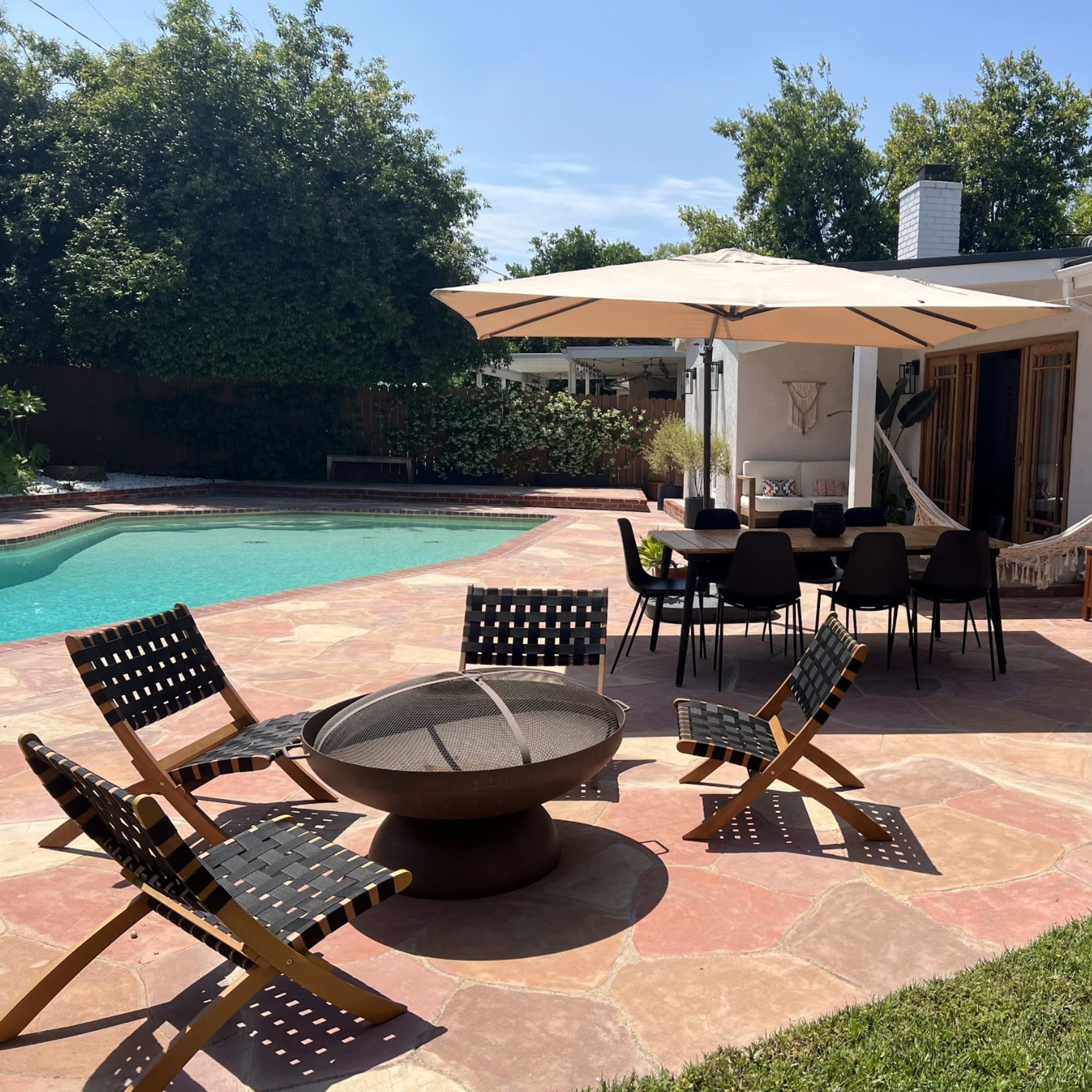 A patio featuring a dining table with chairs, a fire pit, and a hammock near a swimming pool surrounded by greenery.