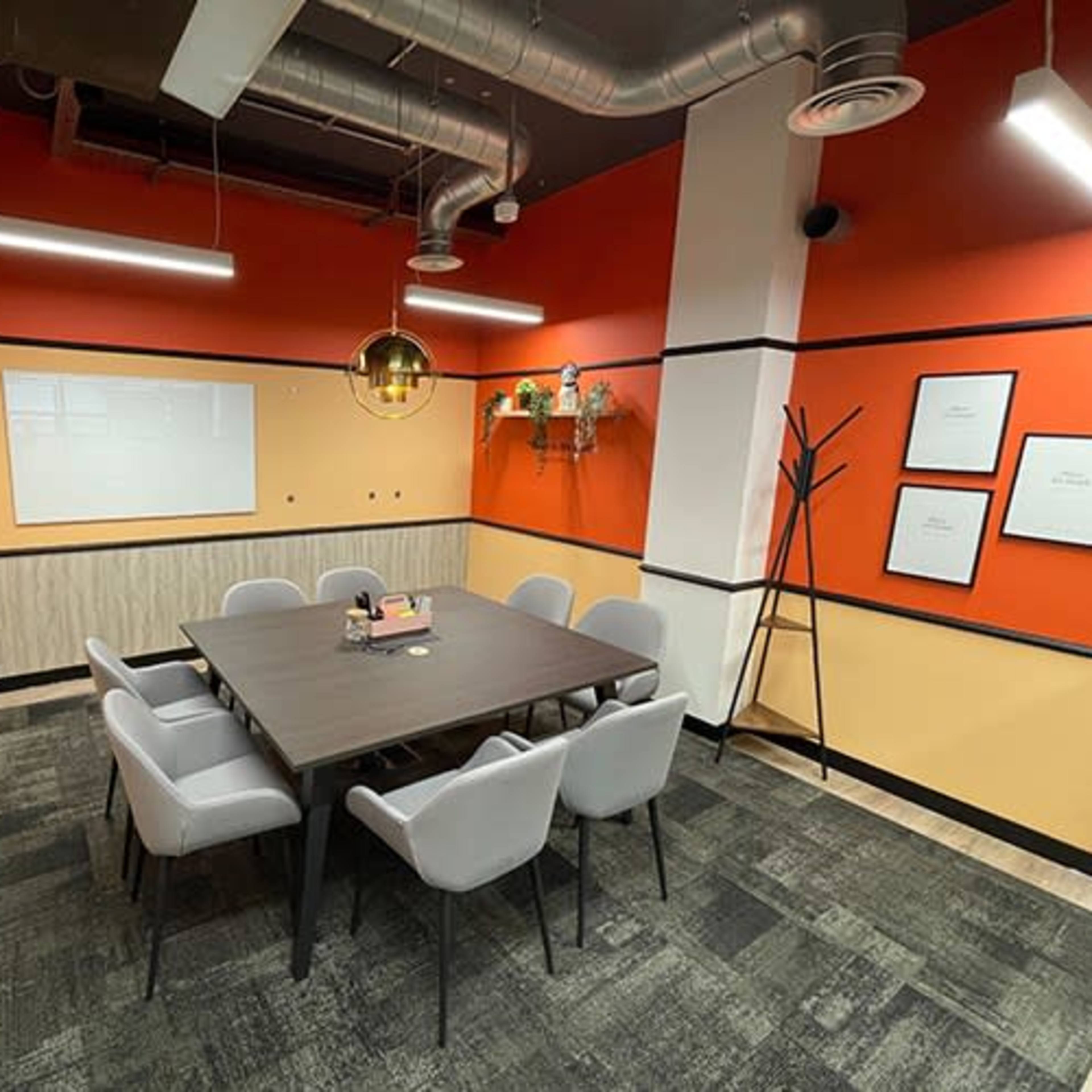 A modern meeting room with a large table surrounded by eight chairs, accented by orange and beige walls and decorative plants.