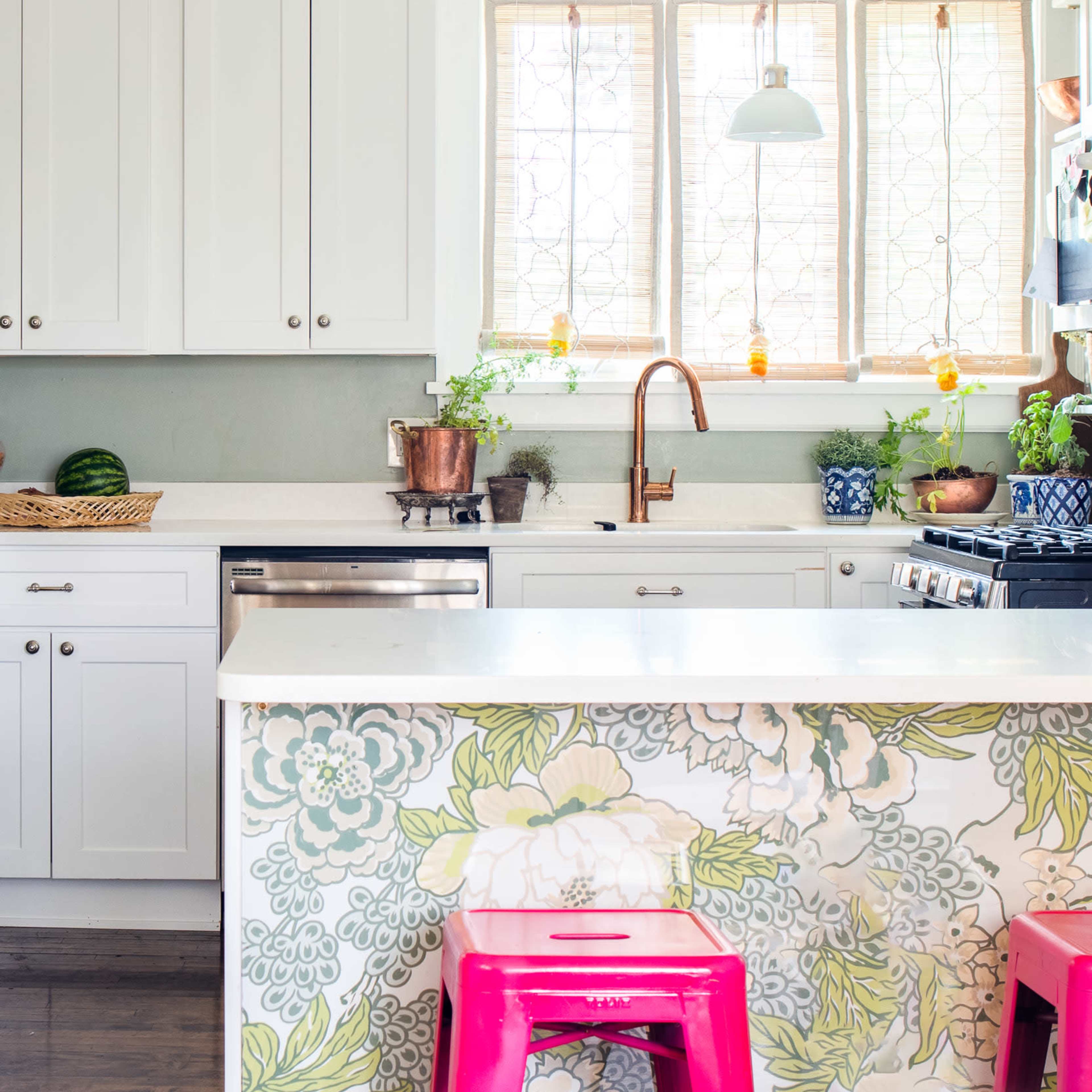 A bright kitchen featuring white cabinetry, a marble countertop, and floral wallpaper, with a pink stool and various plants arranged on the counters.