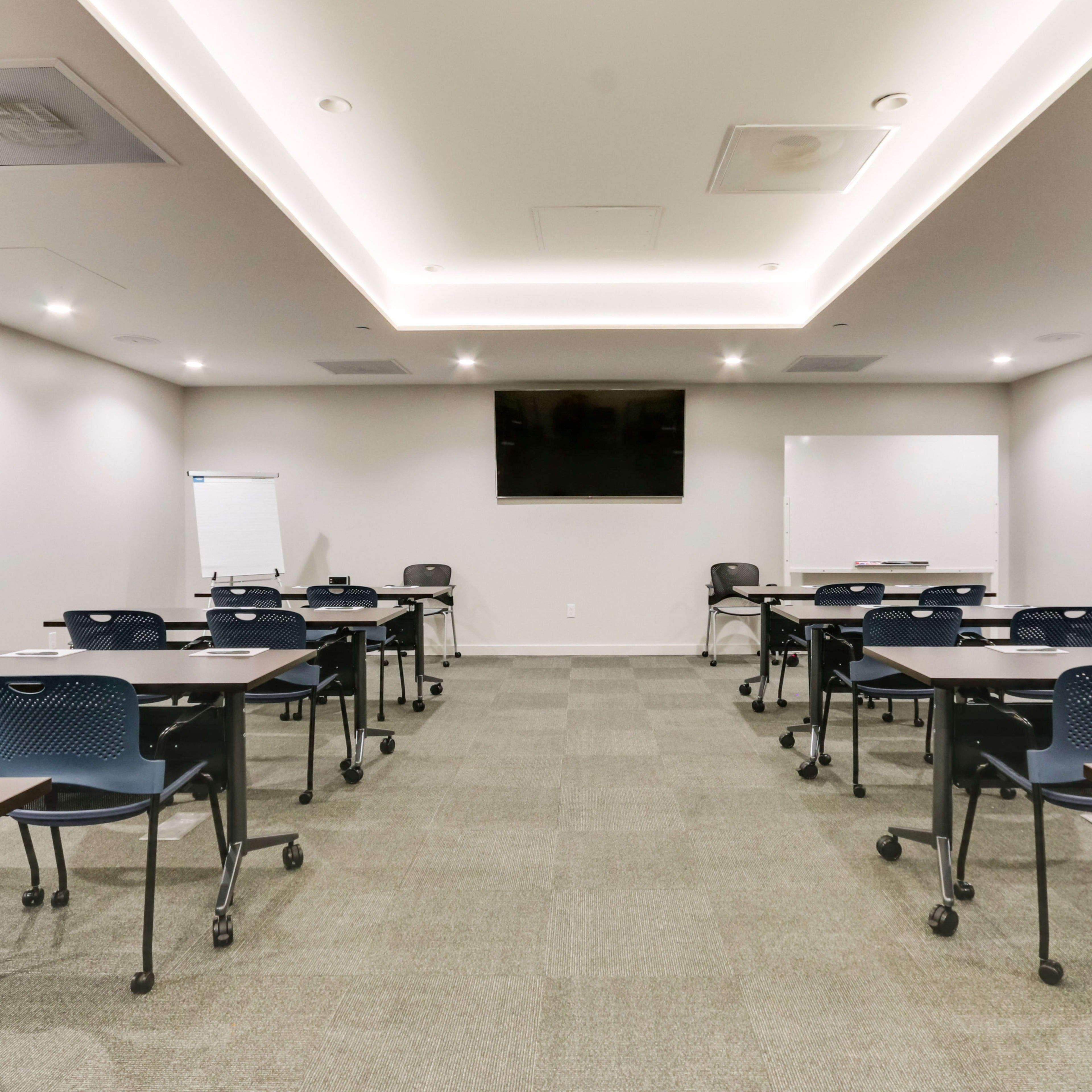 A modern conference room featuring several rows of tables and chairs, a television screen on the wall, and a whiteboard.