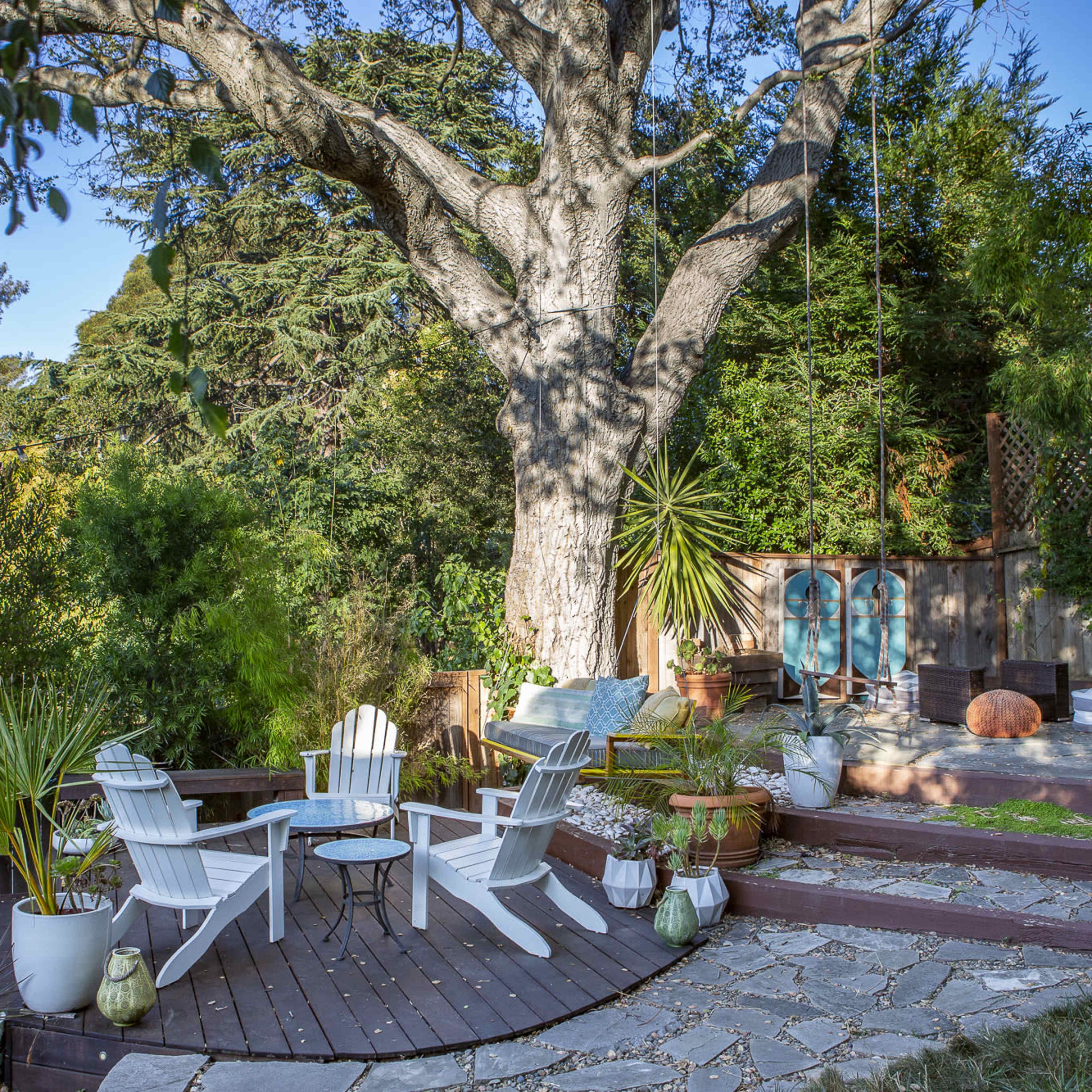 A relaxing outdoor space with white Adirondack chairs arranged around a stone patio, a large tree in the background, and a variety of potted plants.