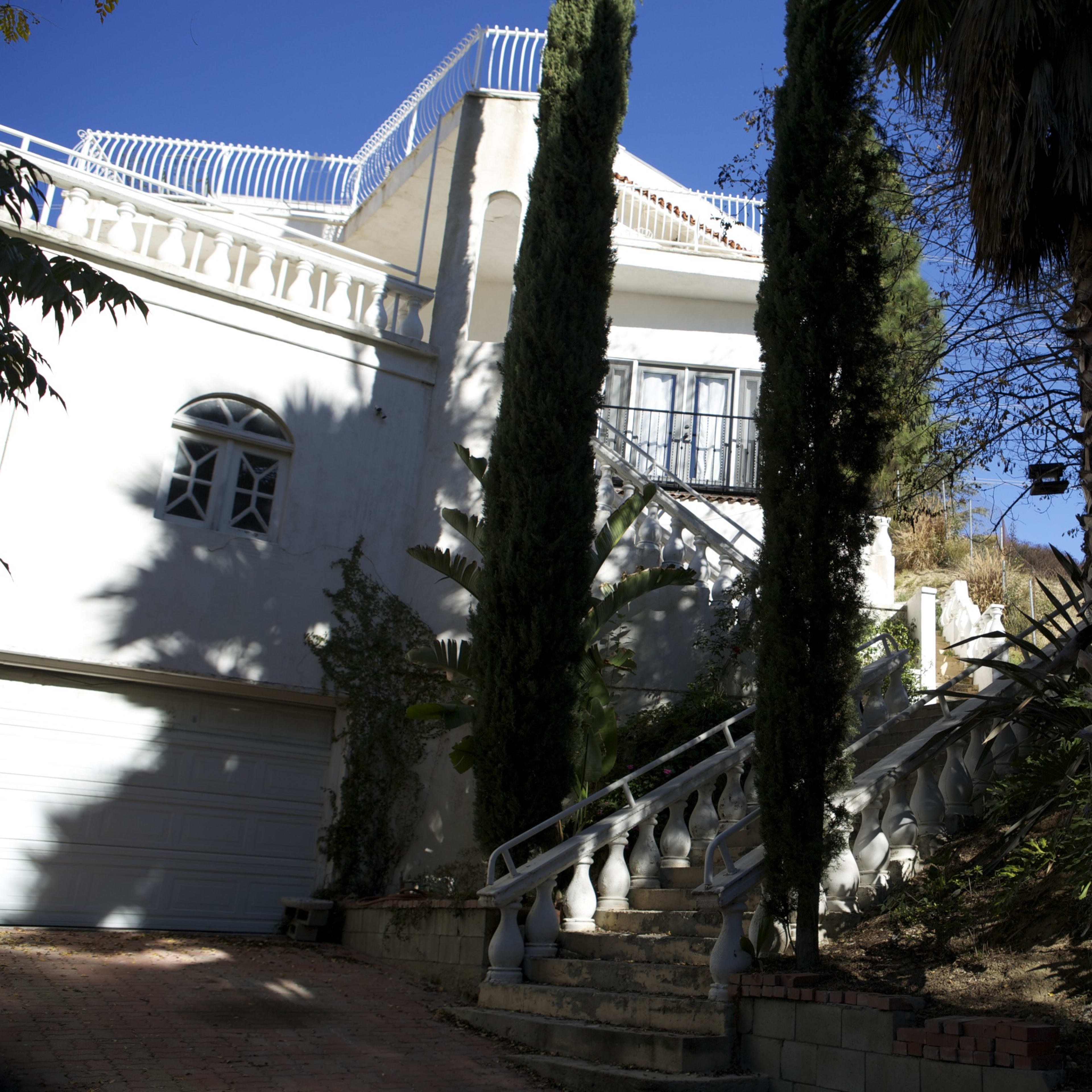 A white house with a balcony and stairs flanked by palm trees, set against a clear blue sky.